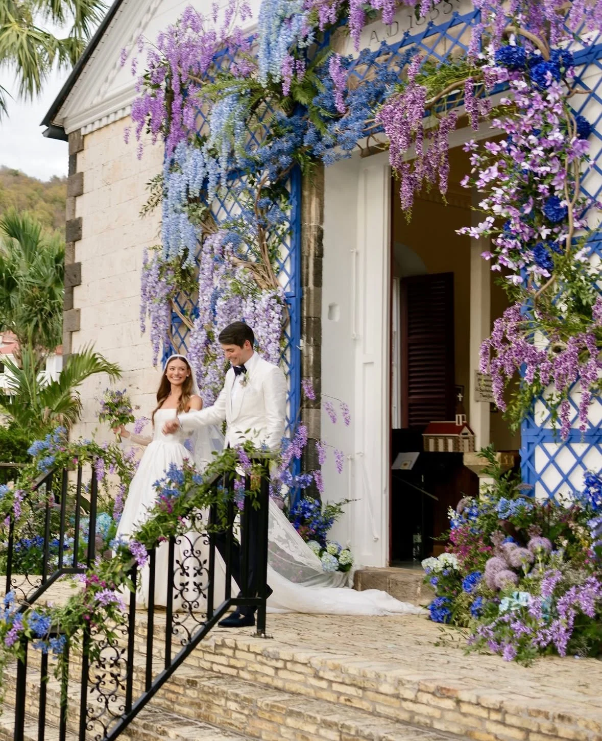 Un couple en robe de mariage se tenant la main devant une bâtisse décorée de guirlandes de fleurs violettes et bleues.