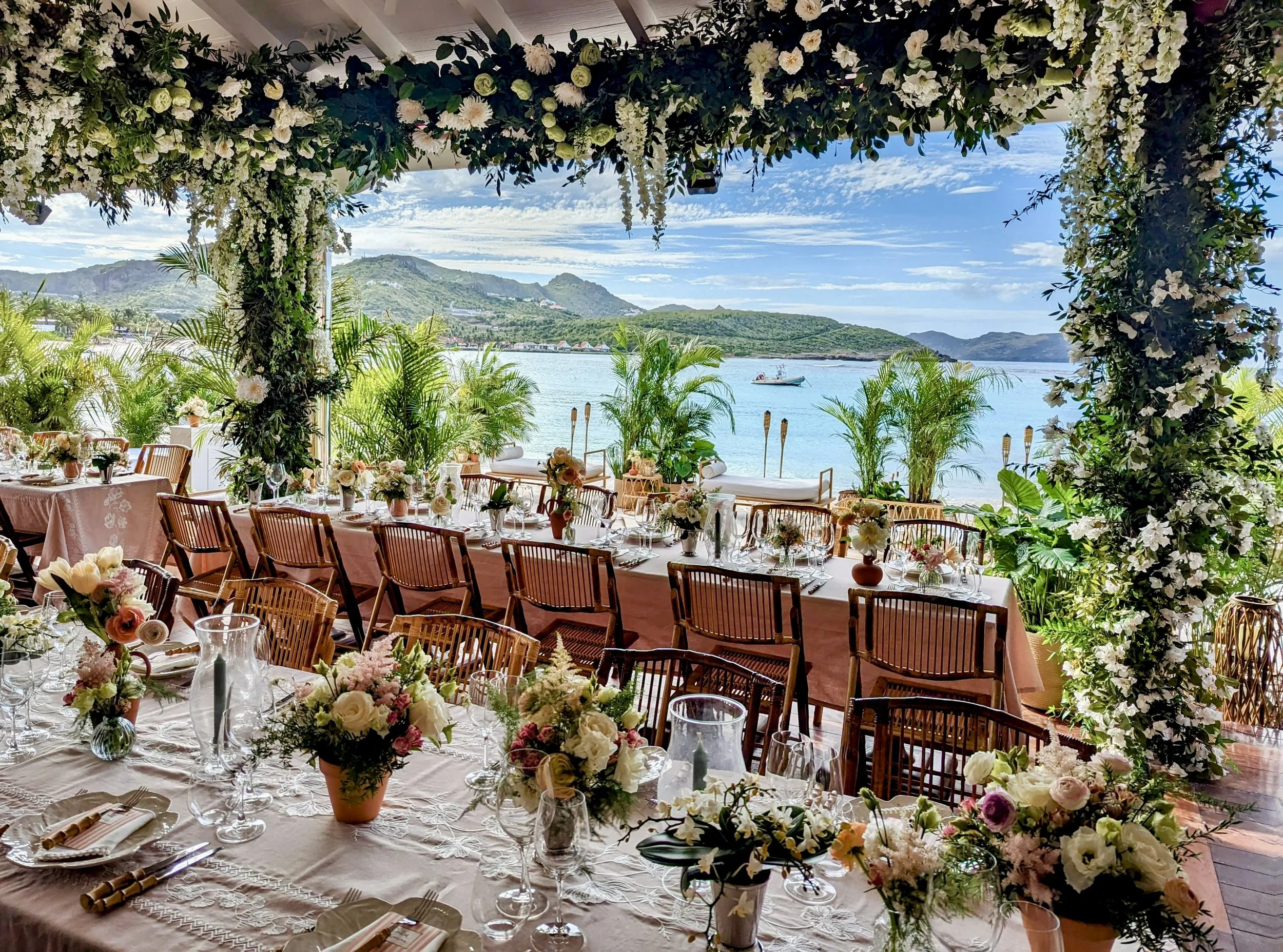 Vue d'une salle de réception décorée pour un mariage en plein air, avec vue sur la mer, des tables avec des arrangements floraux et des chaises en bois.
