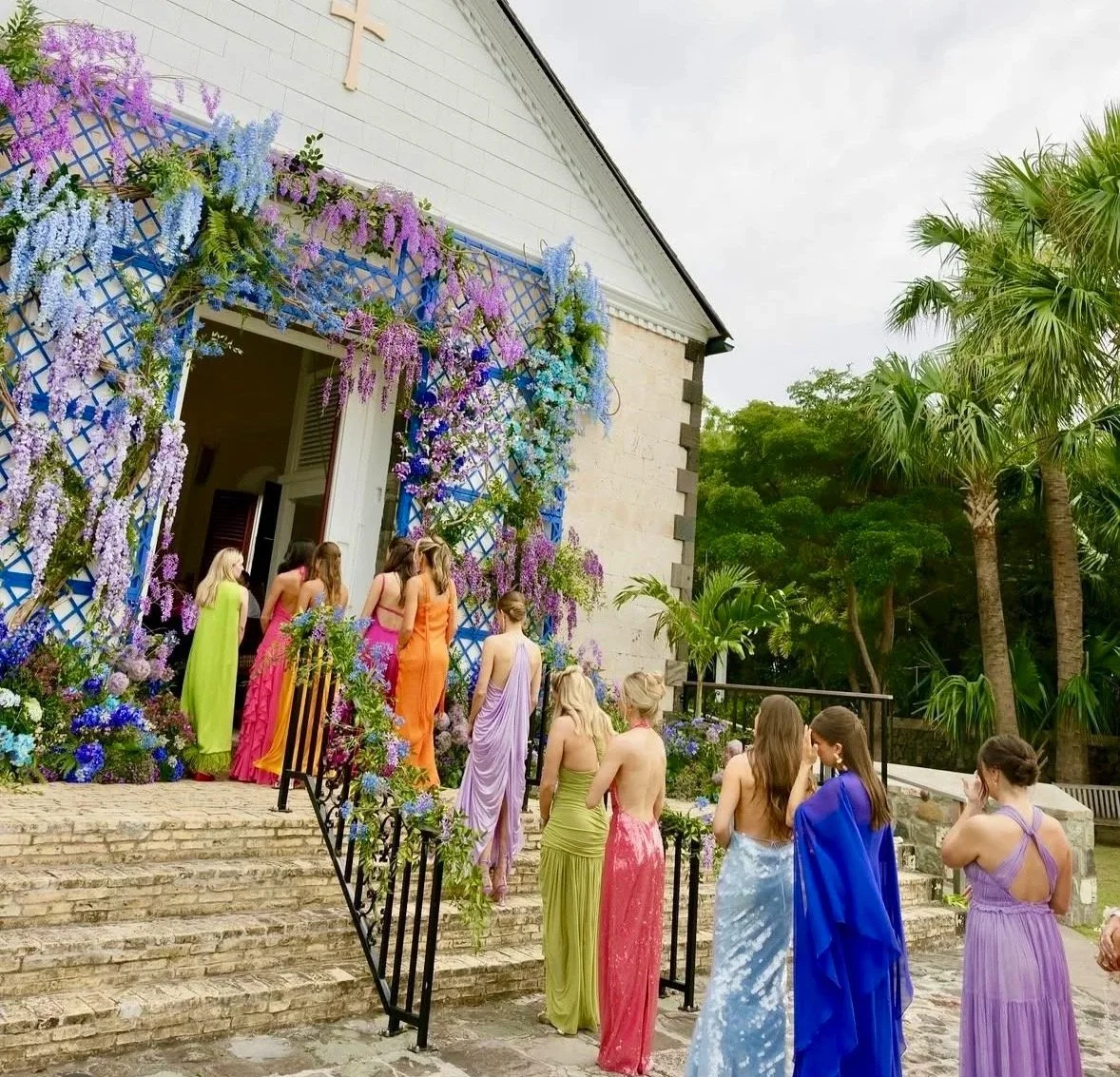 Groupe de jeunes femmes habillées en robes longues colorées, près de l'entrée d'une chapelle décorée de fleurs violettes et bleues, dans un environnement tropical avec des palmiers.