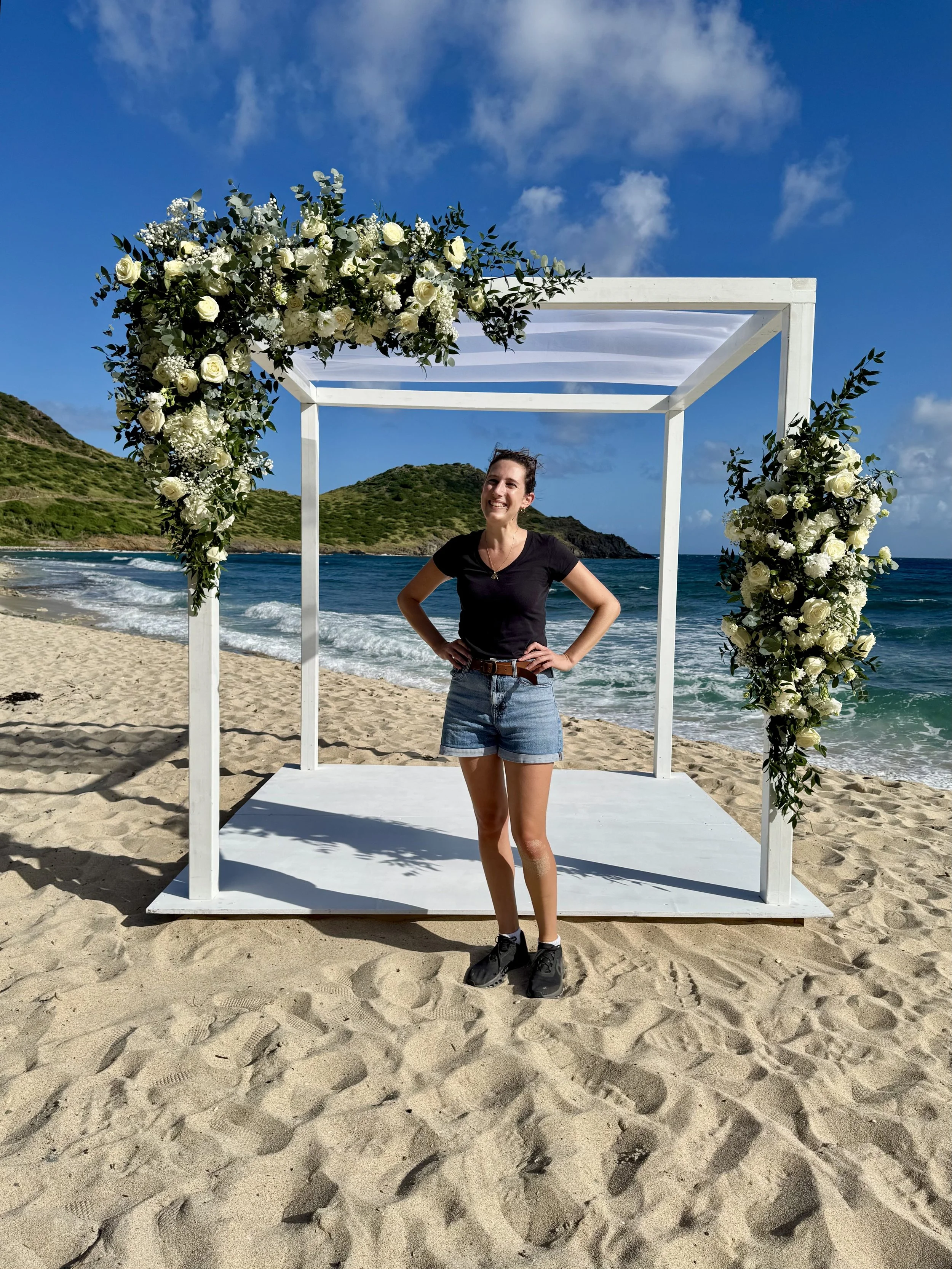 Une femme souriante en t-shirt noir, shorts en jean et baskets, se tient sous une structure blanche décorée de fleurs blanches sur une plage de sable, avec la mer et une colline en arrière-plan, sous un ciel bleu avec quelques nuages.
