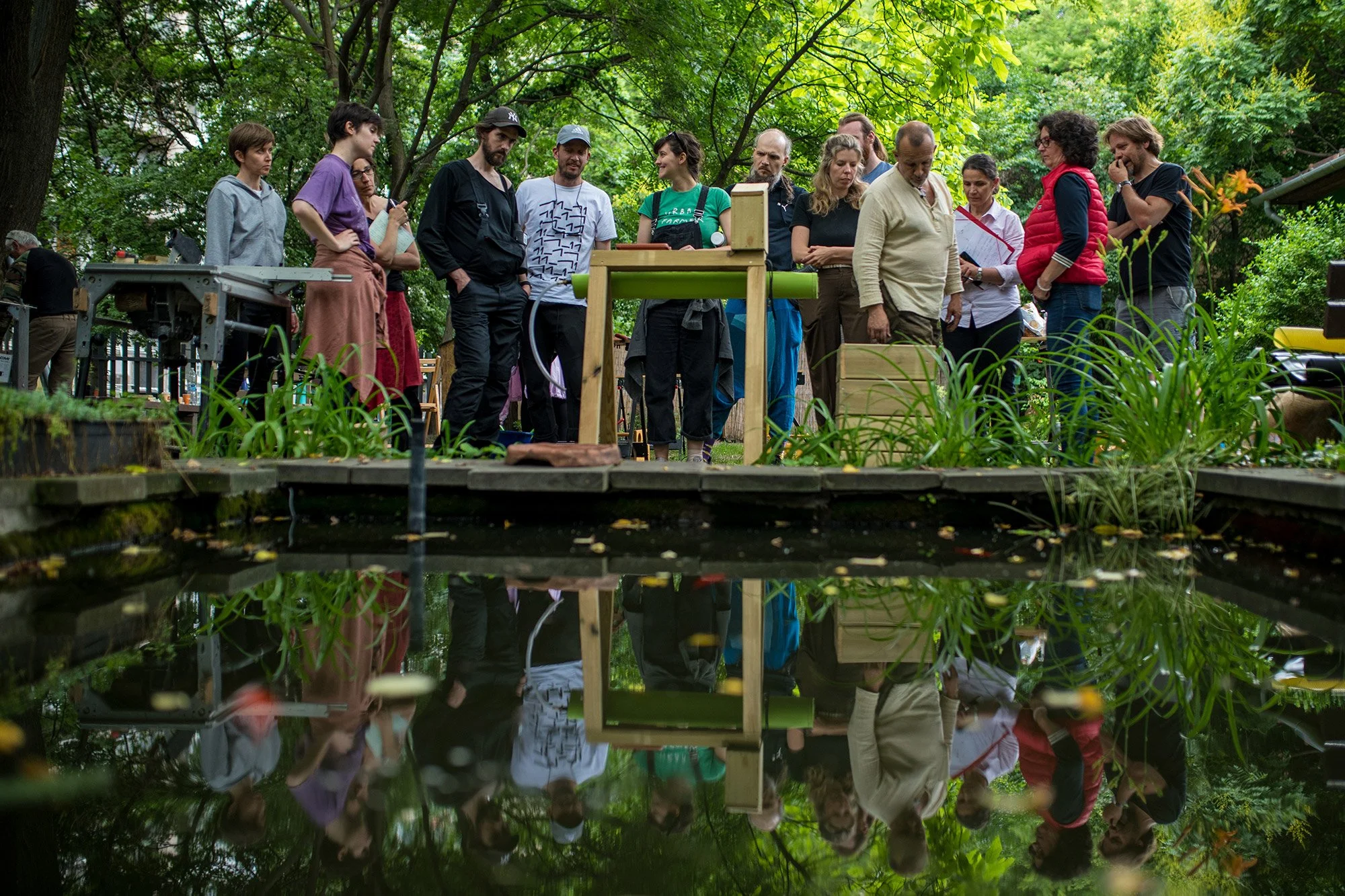 Group of people gathered outdoors around plants and a water feature, engaging in a discussion or demonstration in a lush green park. Permaculture, Permakultúra workshop at HUMUSZ kert, Bartók-negyed, Bartók-Quarter, 11_11.