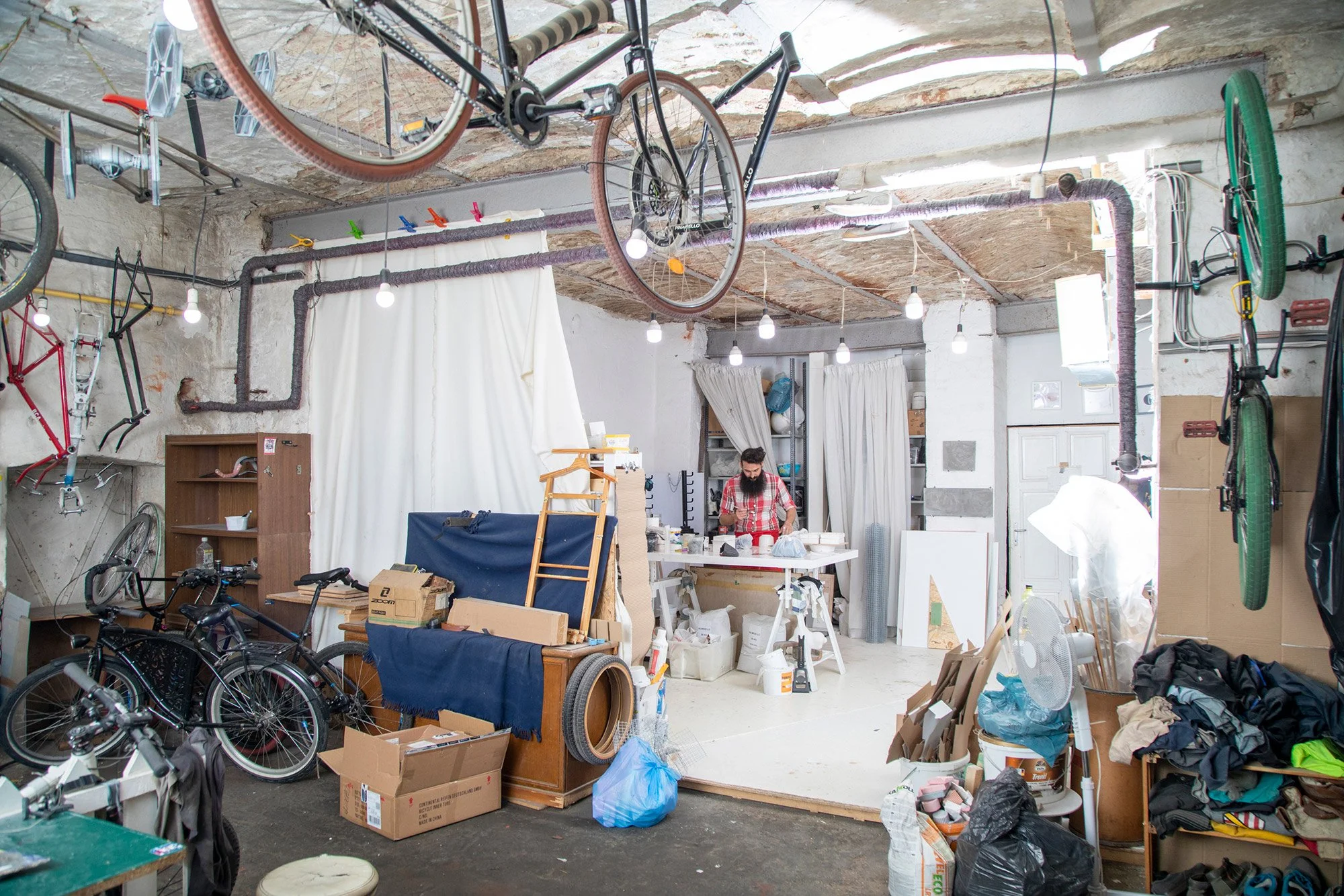 A cluttered workshop or studio with bicycles hanging from the ceiling, a man working at a table, and various tools, boxes, and materials scattered around.
A studio of 11_11 at Bartók-Quarter. Bartók-negyed műterem.