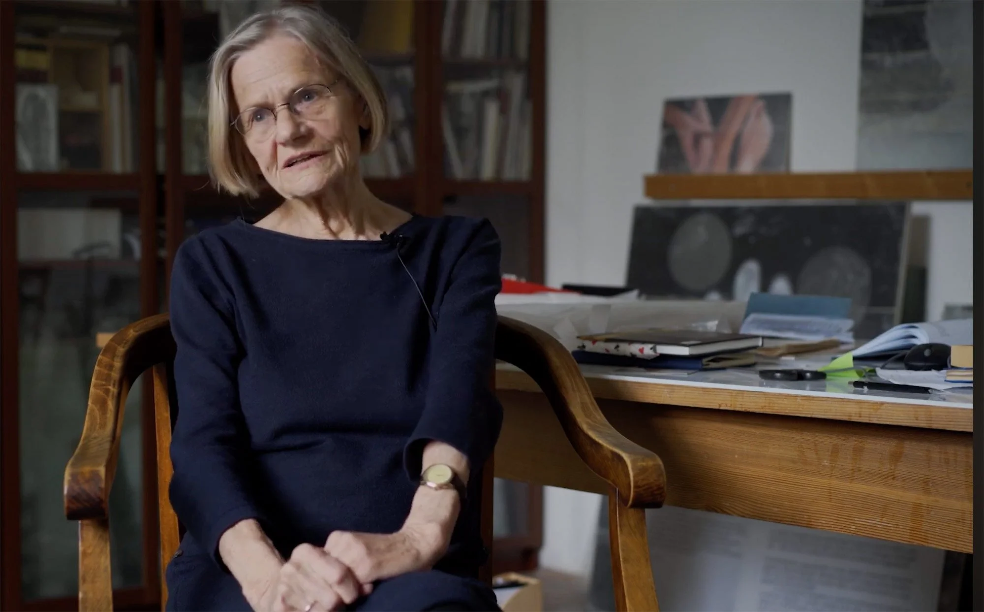 An elderly woman with glasses and shoulder-length gray hair, wearing a black top, sitting in a wood chair in a room with a cluttered desk and bookshelves in the background. Ilona Lovas giving an interview about Velem Textile Workshop.