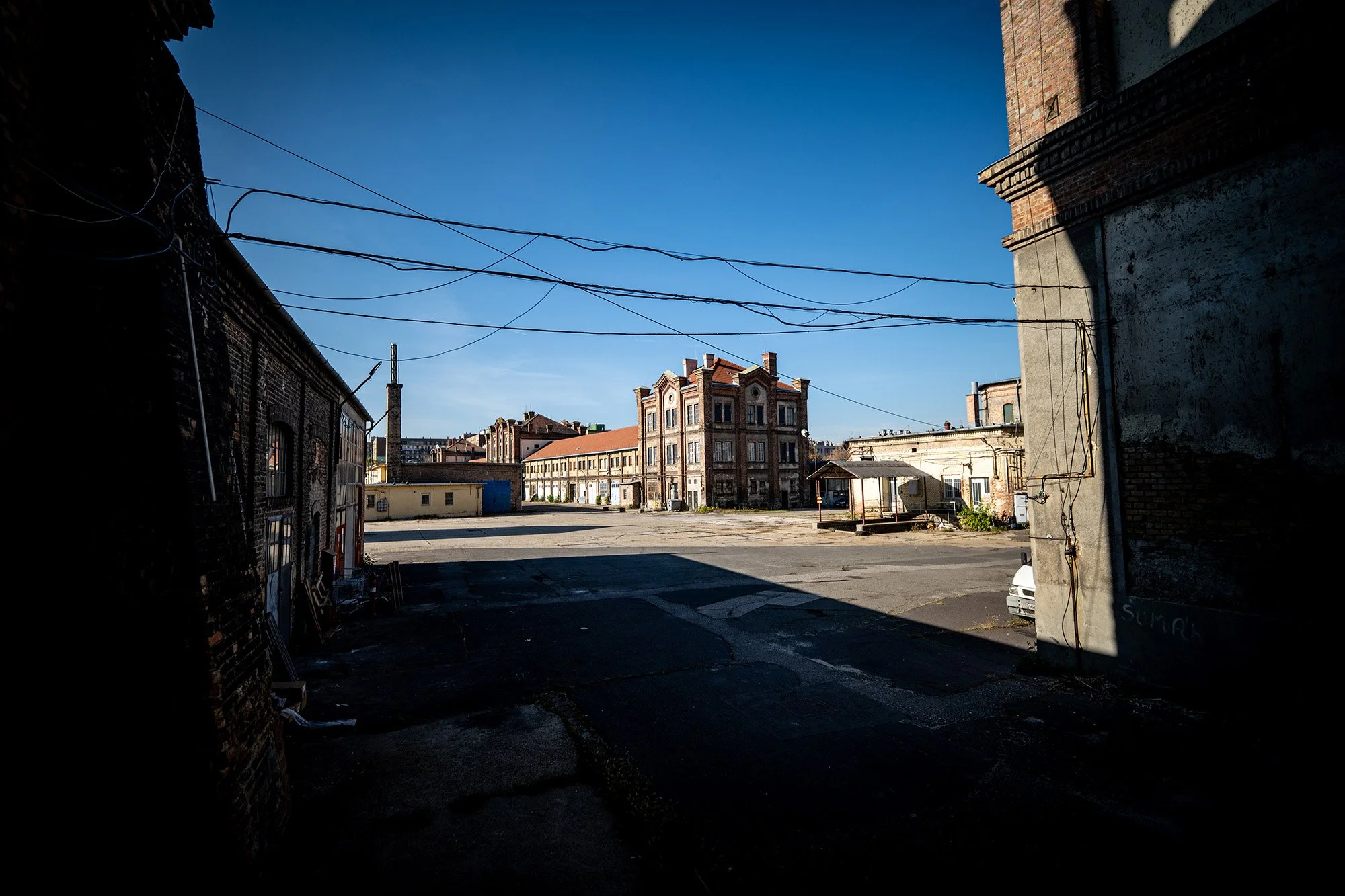An empty urban lot surrounded by old brick and concrete buildings under a clear blue sky, with overhead power lines. S1 Kőbánya, Budapest, former brewery site, postindustrial site.