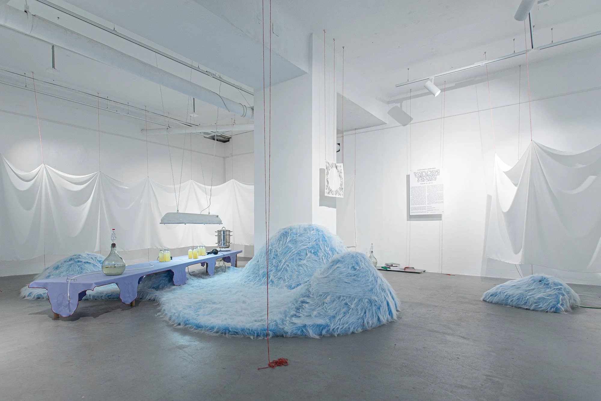 An art installation by Fuzzy Earth at 1111 Gallery and Community Space featuring a white room with draped white fabric, a purple table with glass bottles, a large fluffy blue rug resembling clouds. At Bartók-negyed, Bartók-Quarter.