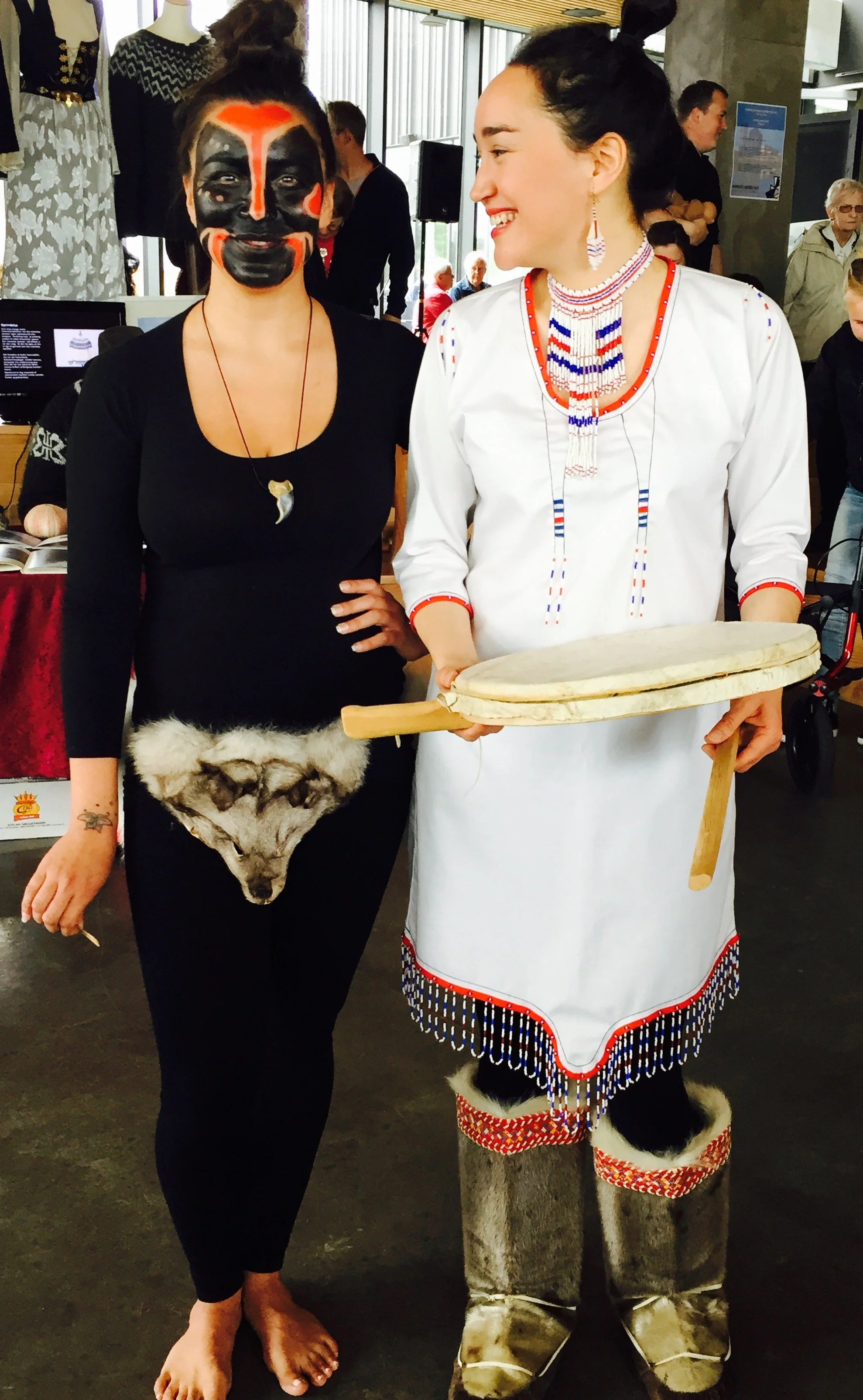 To kvinder i traditionel grønlandsk dragt ved et kulturelt arrangement. Grønlandsk maskedanser, mens den anden holder en grønlandsk tromme til grønlandsk maskedans og inuit kulturtraditioner.