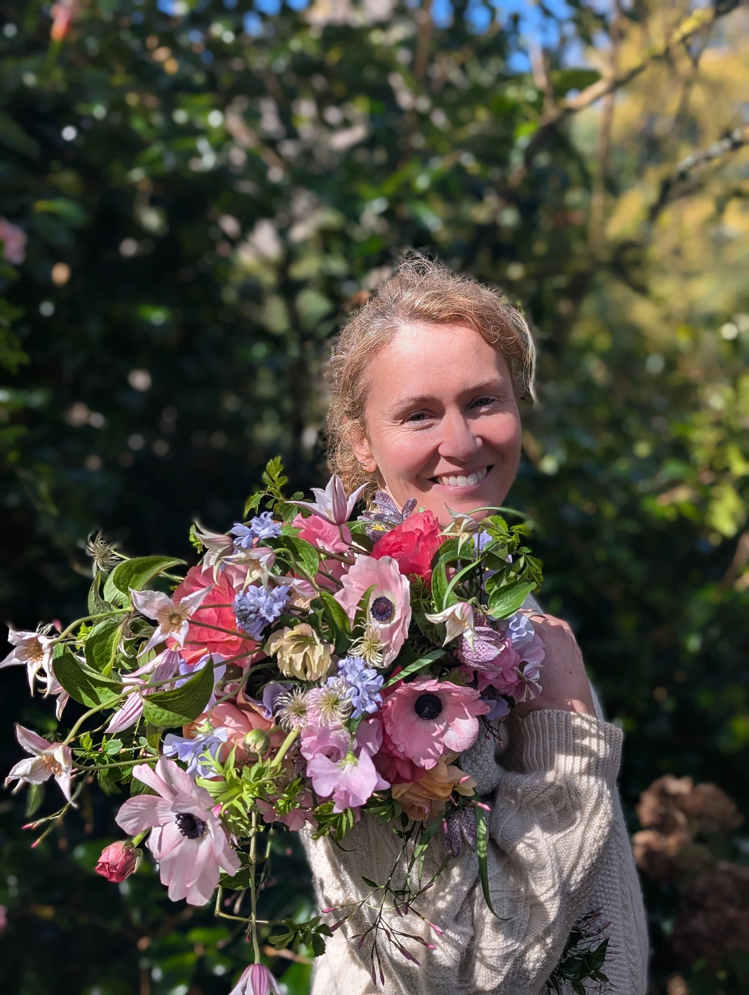 Kat smiling and holding a colorful bouquet of flowers outdoors with green foliage background.