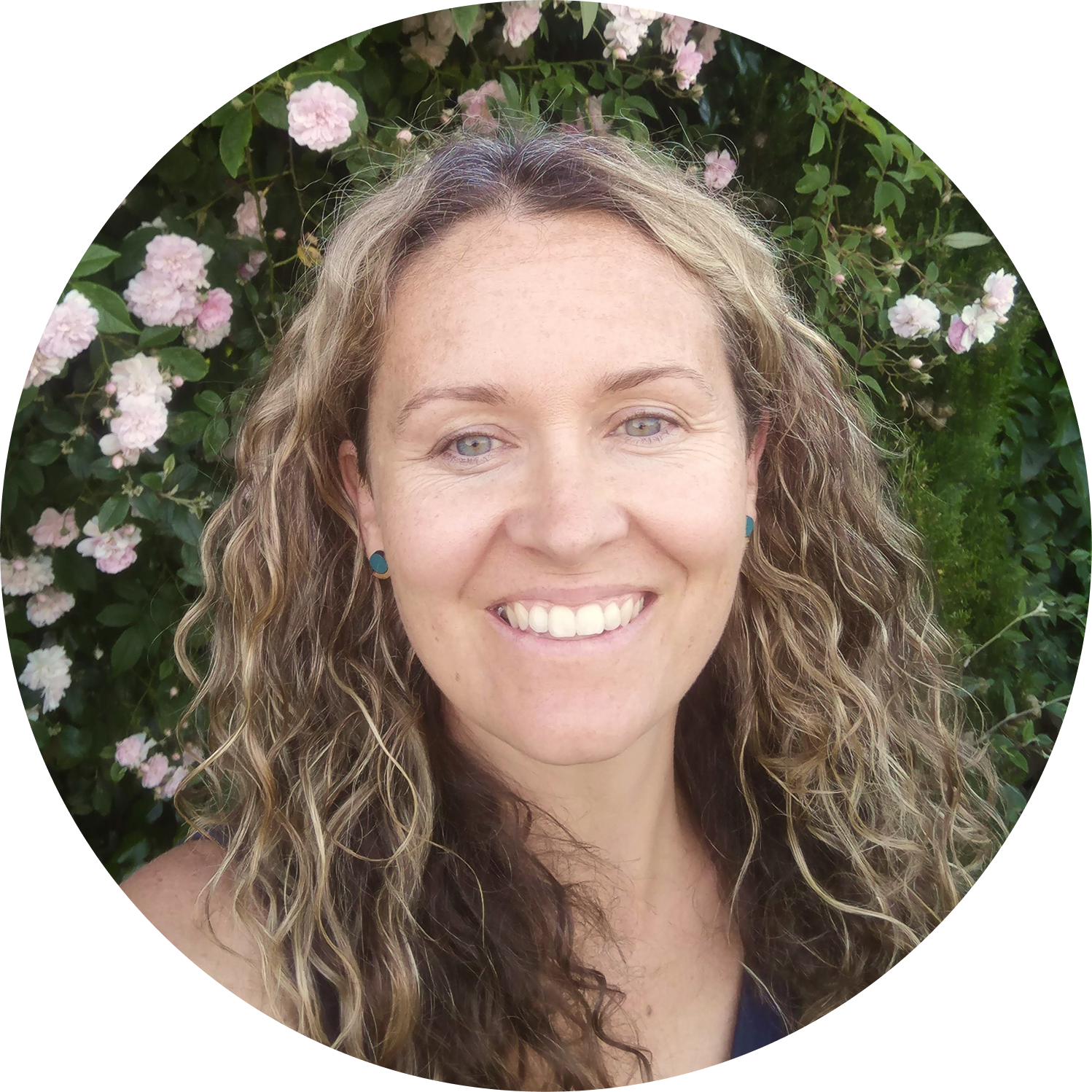 Kat, a smiling woman with curly hair wearing blue earrings, outdoors in front of a pink rose bush