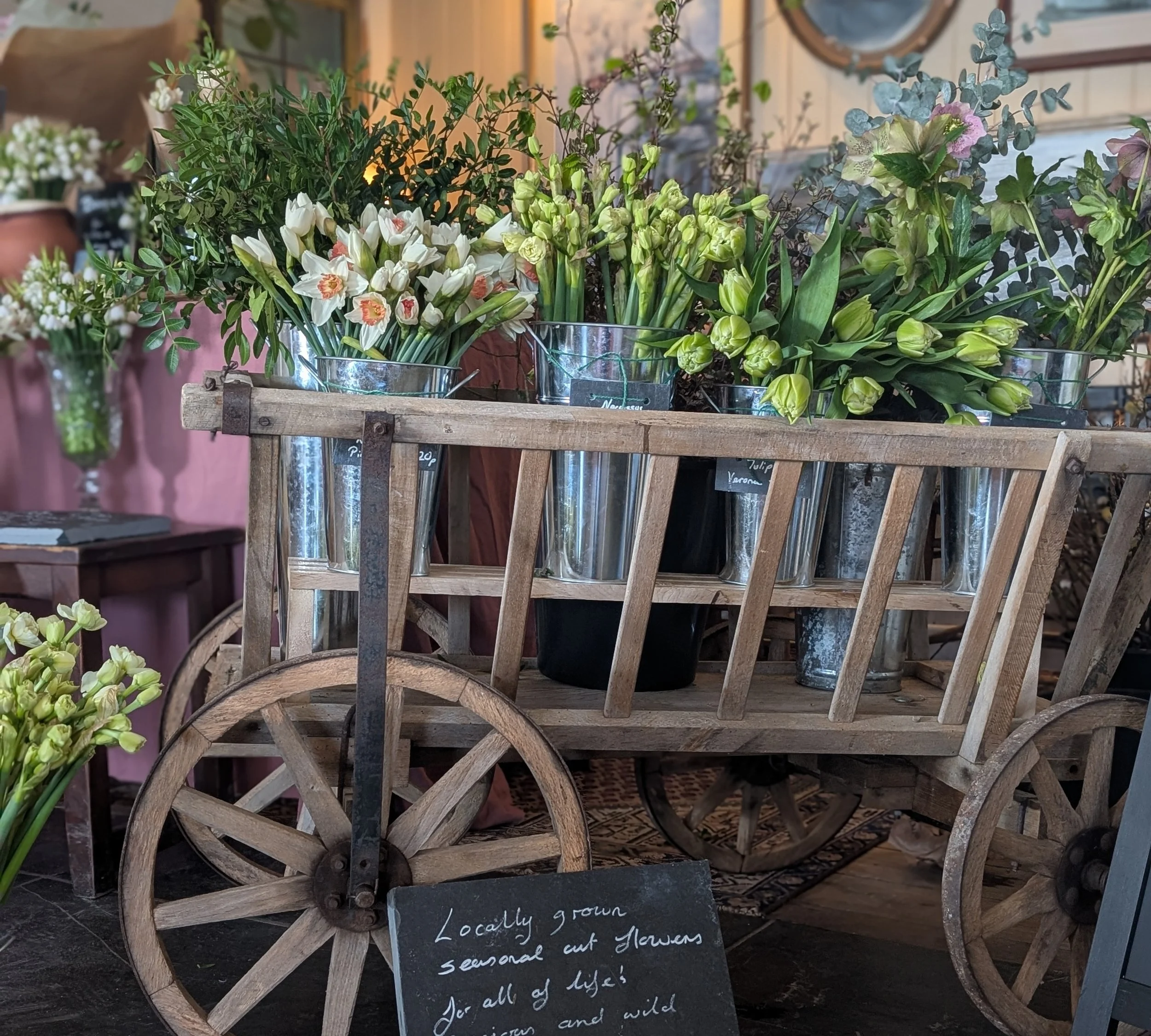 A wooden  Victorian cart filled with cut flowers in metal buckets, including white, pale green, and yellow flowers, displayed in a floral shop.