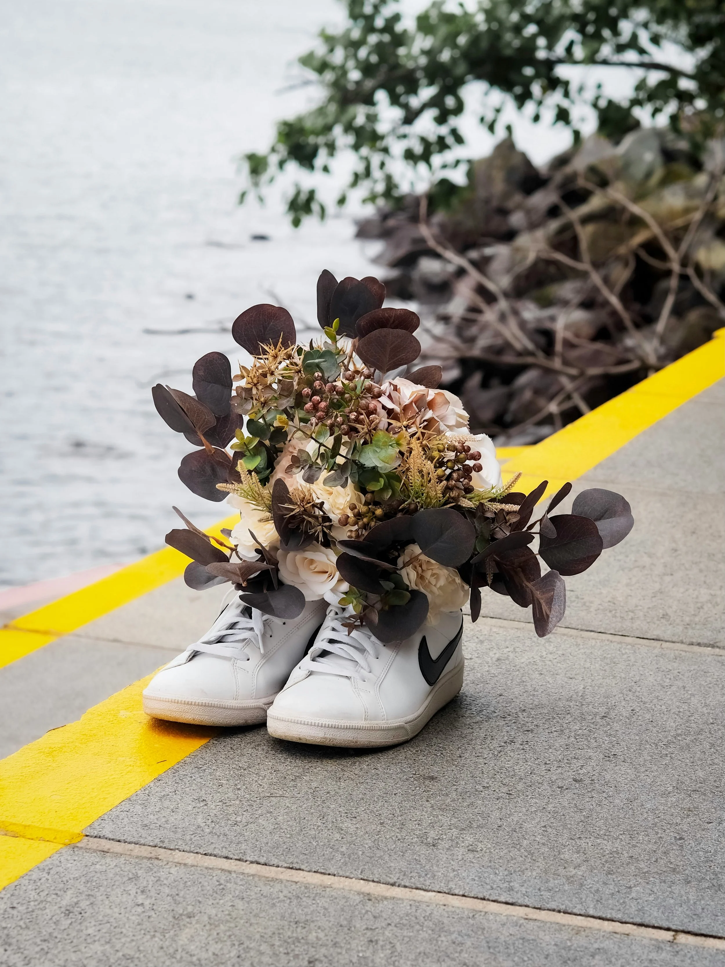 White Nike sneakers with a bouquet of brown, white, and green flowers on top, placed on a concrete sidewalk near water, with trees in the background.