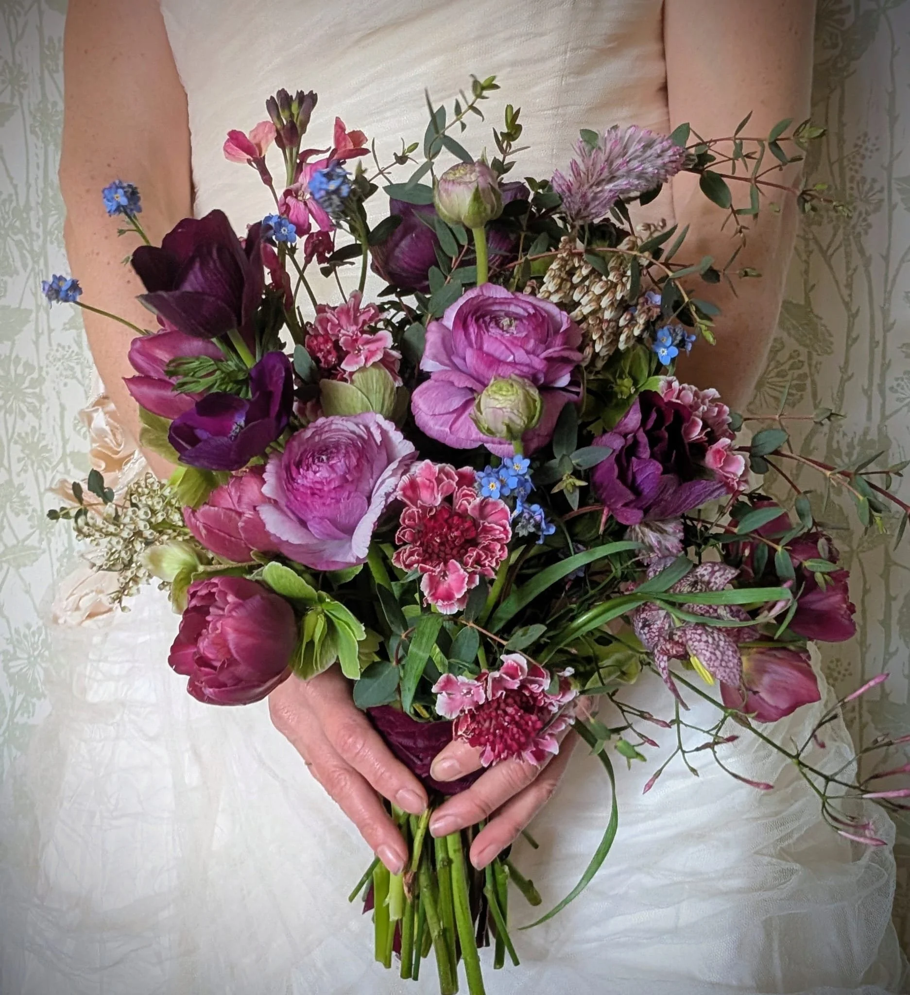 A bride holding a large bouquet of ranunculus, clematis, and tulip flowers with green foliage, in front of a light-colored, patterned background.