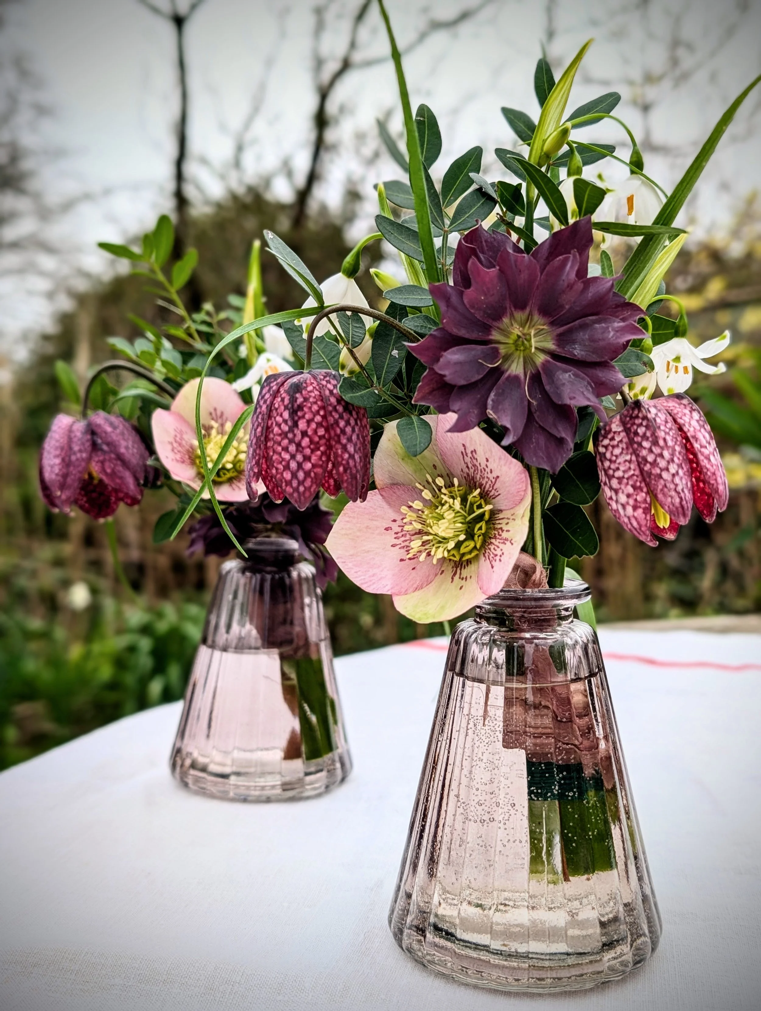 A wedding table arrangement in two pink glass bud vases on a white tablecloth outdoors, featuring pink, purple, and white flowers with green leaves.
