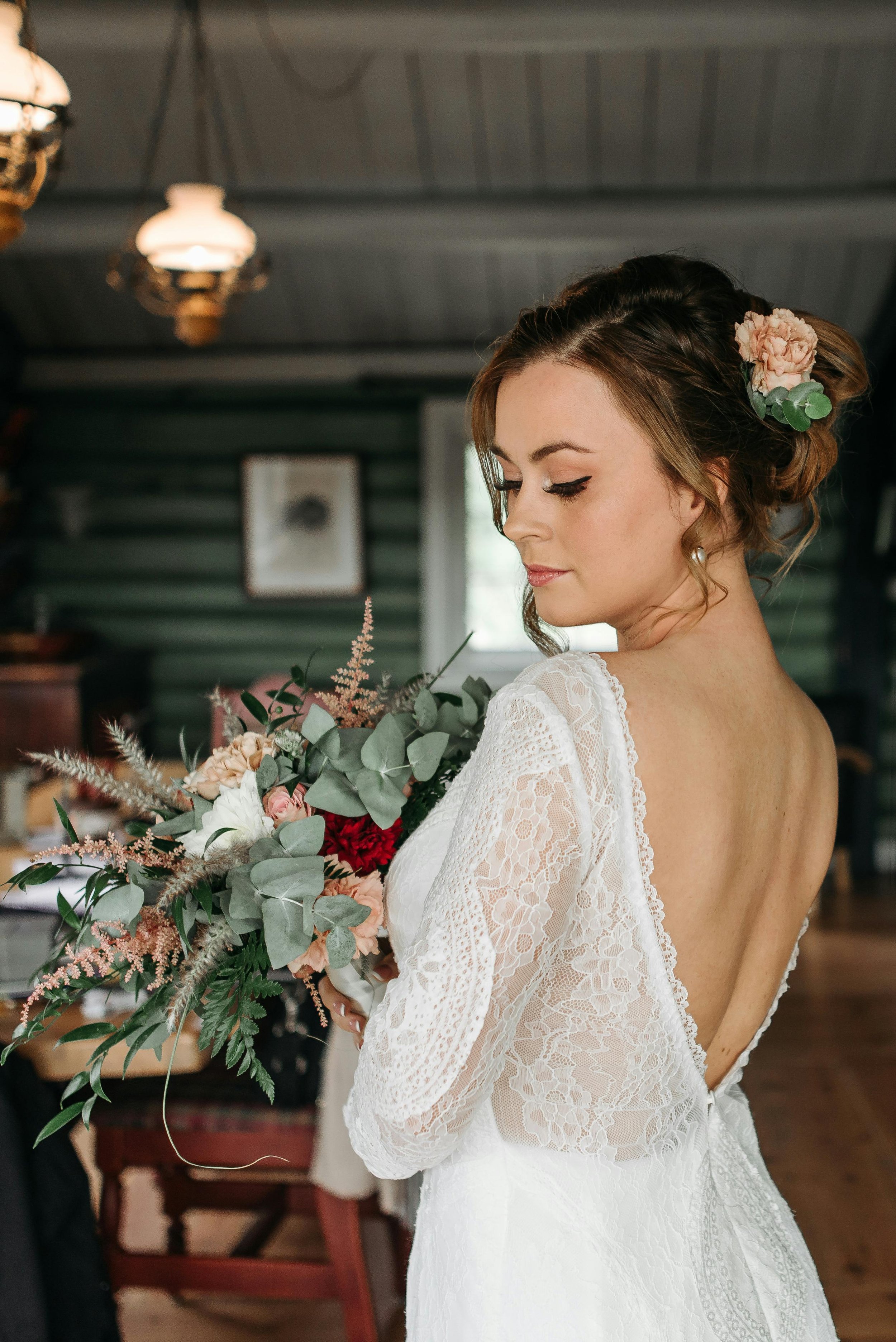 A bride in a lace wedding dress holding a bouquet of flowers, with a flower in her hair, in a rustic indoor setting.