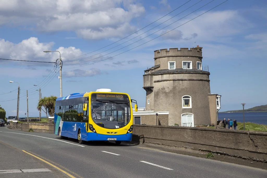 A modern blue and yellow bus on a coastal road near a round castle-like building with a view of the water and a cloudy sky in the background.