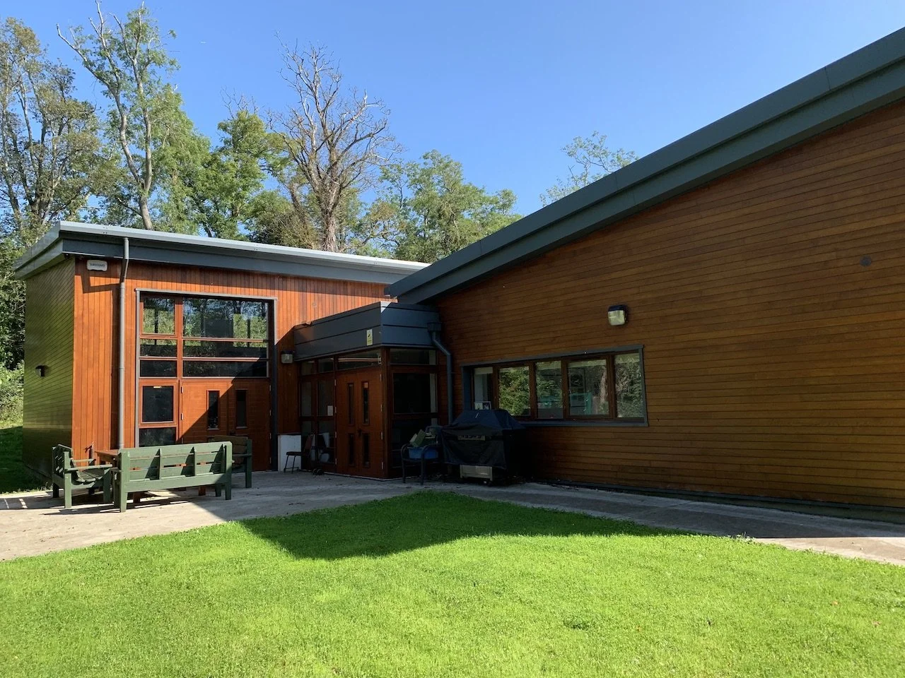 A modern house with wood siding and large windows, with outdoor seating and a barbecue grill on a patio, surrounded by green grass and trees under a blue sky.