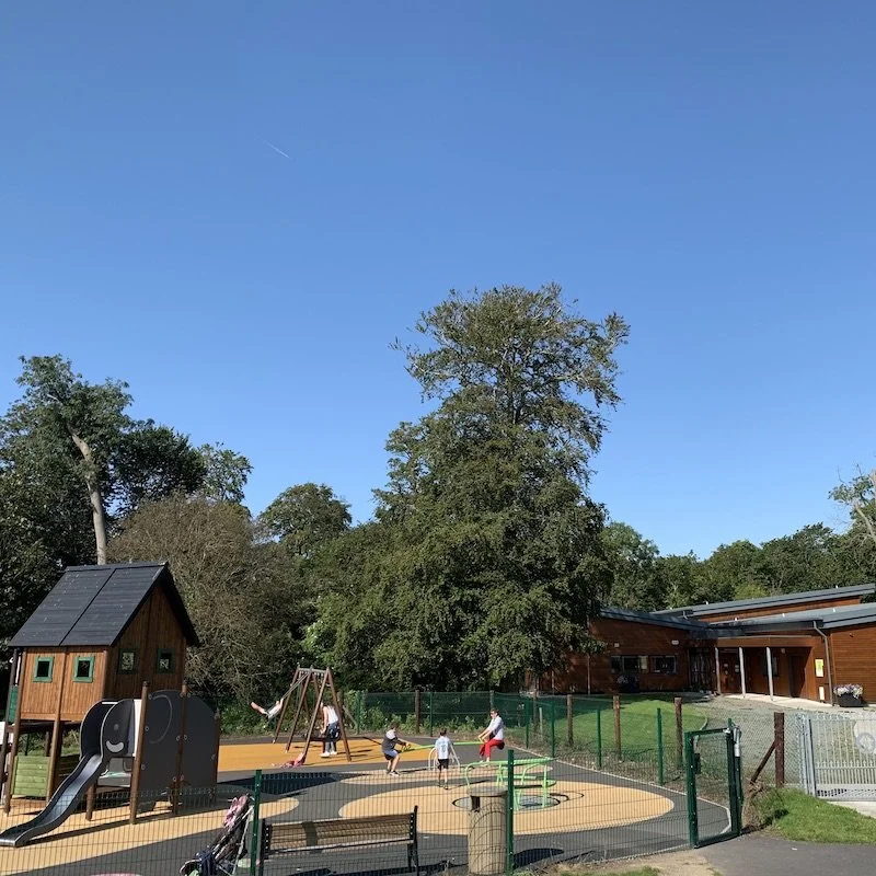 A playground with swings, a slide, and a playhouse, surrounded by a fence and trees, on a sunny day.