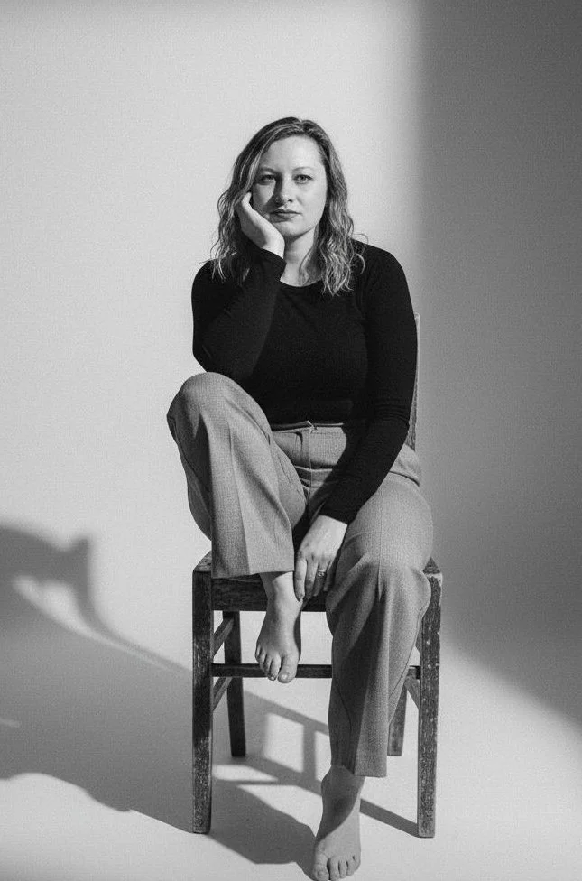 Black-and-white photo of a woman sitting on a wooden chair with one leg crossed over the other. She is resting her head on her hand and looking at the camera with a neutral expression. She has shoulder-length wavy hair and is wearing a long-sleeved black top and light-colored pants.