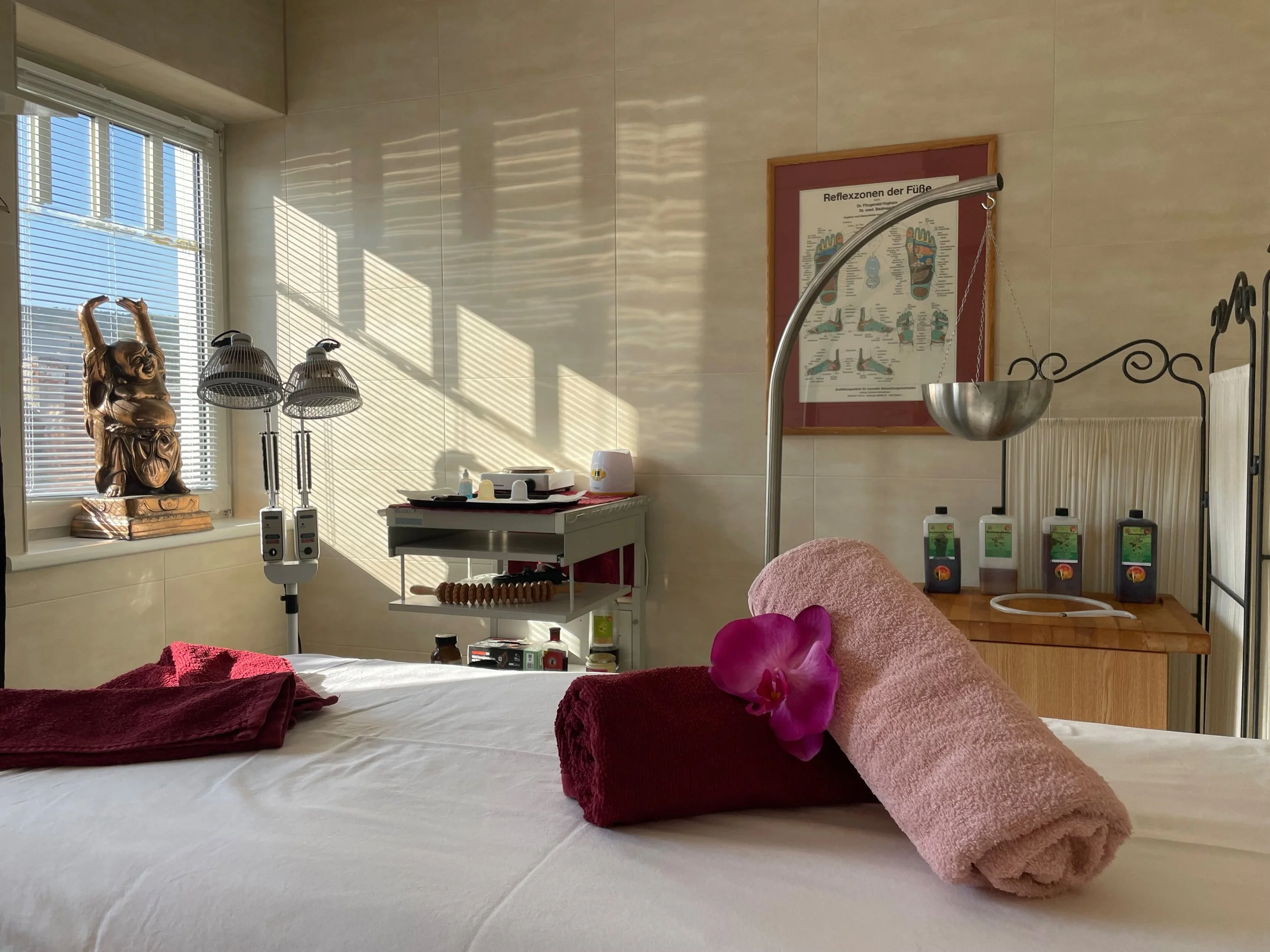 Ein Behandlungsraum mit Therapietisch, eingerollten Handtüchern, einer Orchidee, Skulptur, medizinischer Ausrüstung, Pflanzenölflaschen und Reflexzonenbild an der Wand.