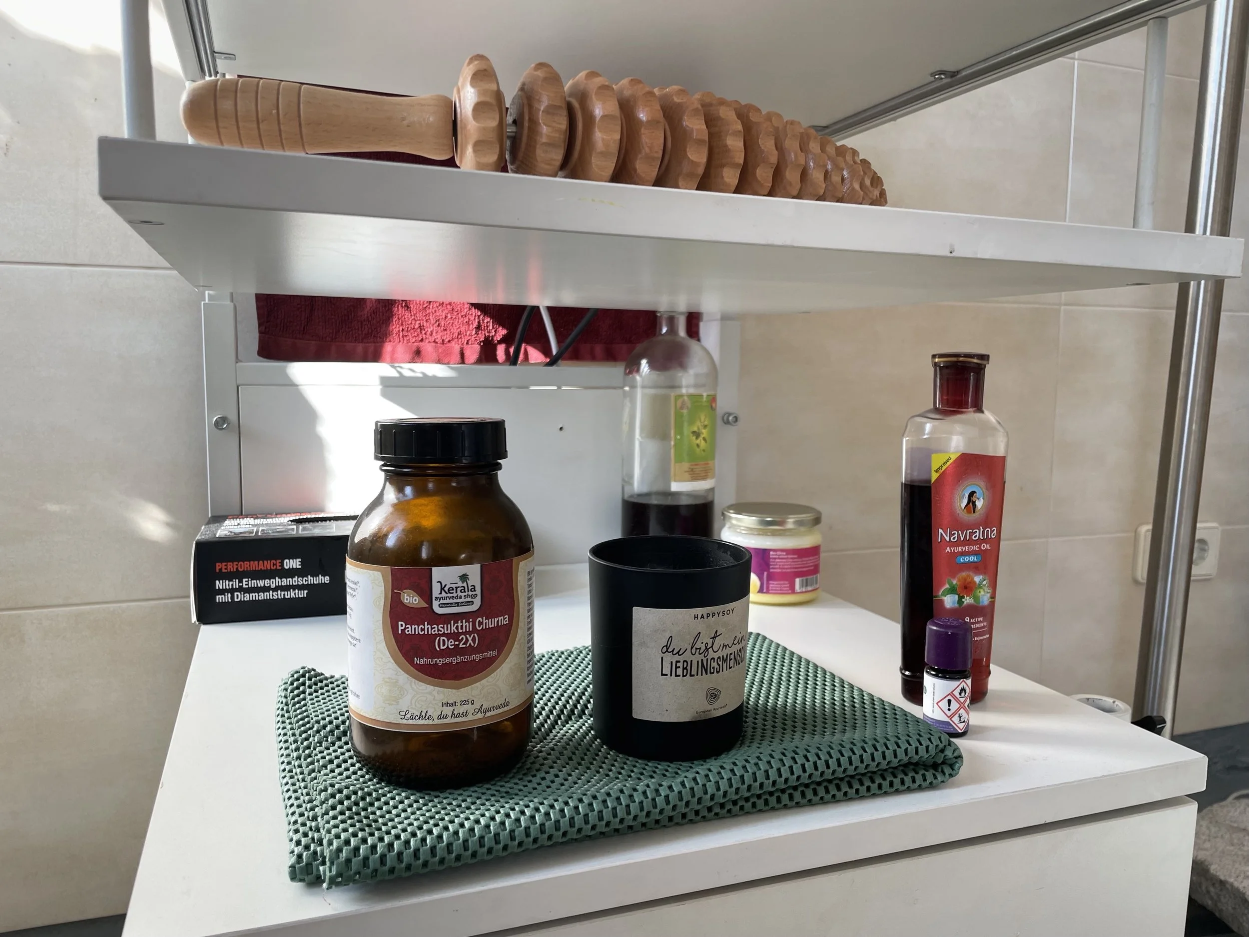 Verschiedene Haushalts- und Gesundheitsprodukte auf einem weißen Regal, darunter ein brauner Behälter mit rotem Deckel, eine Flasche mit schwarzer Flüssigkeit, eine kleine Flasche mit Warnhinweis, ein schwarzer Becher und eine grüne Stoffserviette.