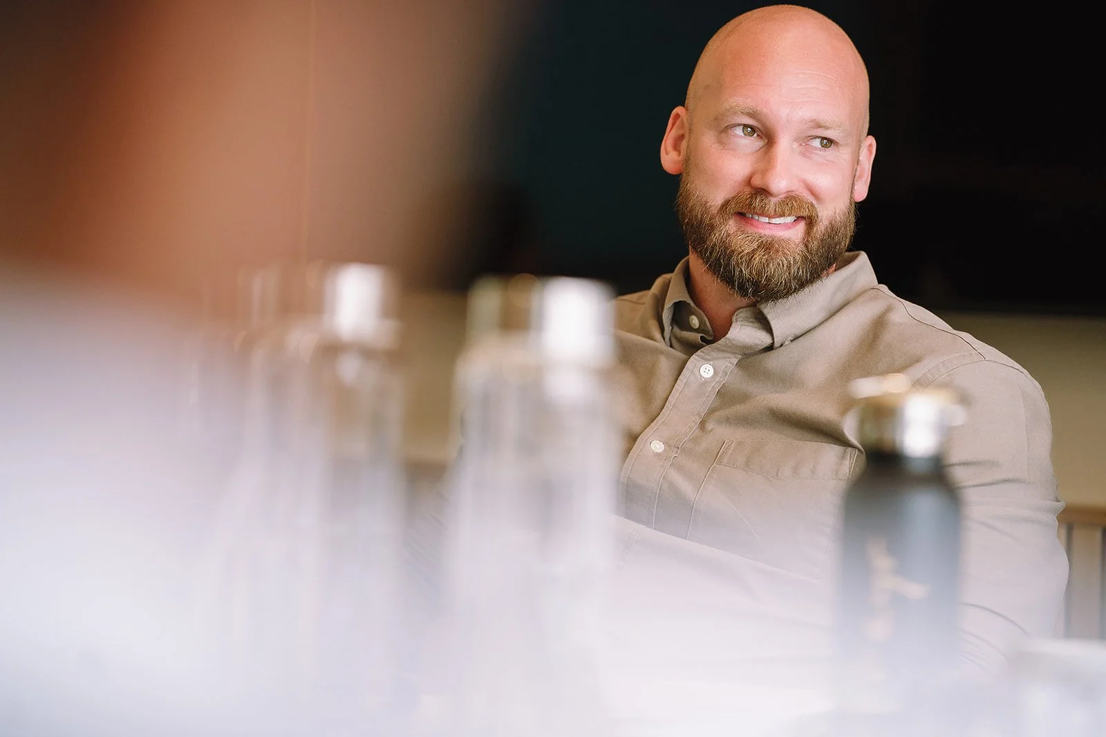 Smiling bald, bearded man in a beige button-down shirt seated behind blurred bottles