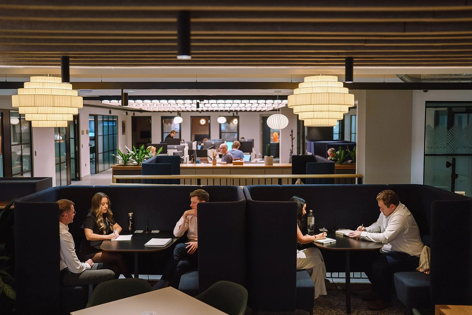 People working and dining in a modern coworking lounge with private booths, plants, and pendant lighting