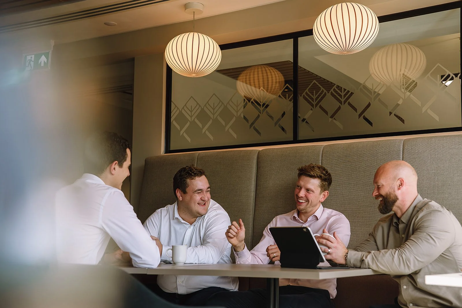 Four coworkers laughing together at a cafe table, with a tablet and coffee cup.