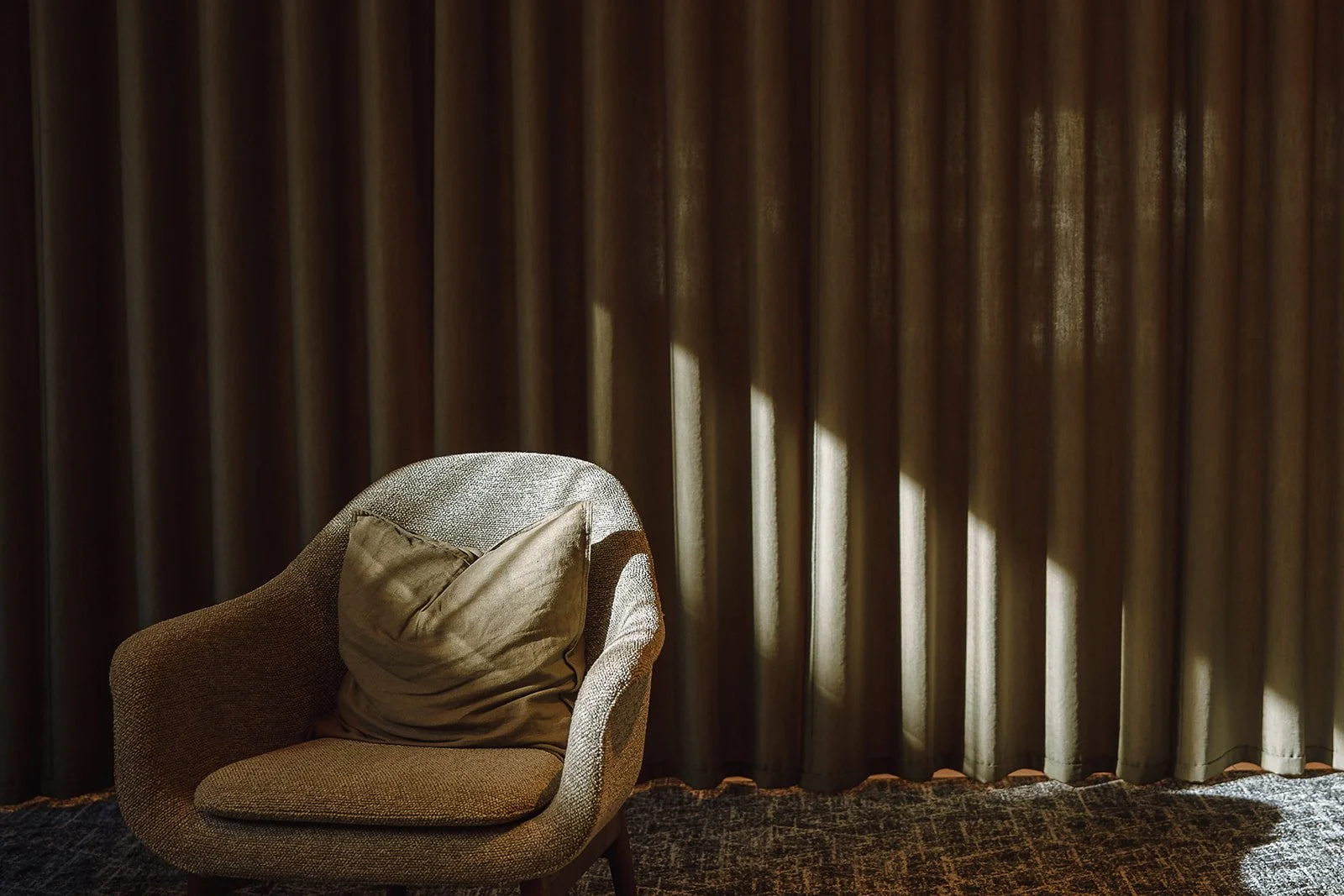 Empty brown armchair with a beige pillow in front of dark curtains with sunlight streaming through