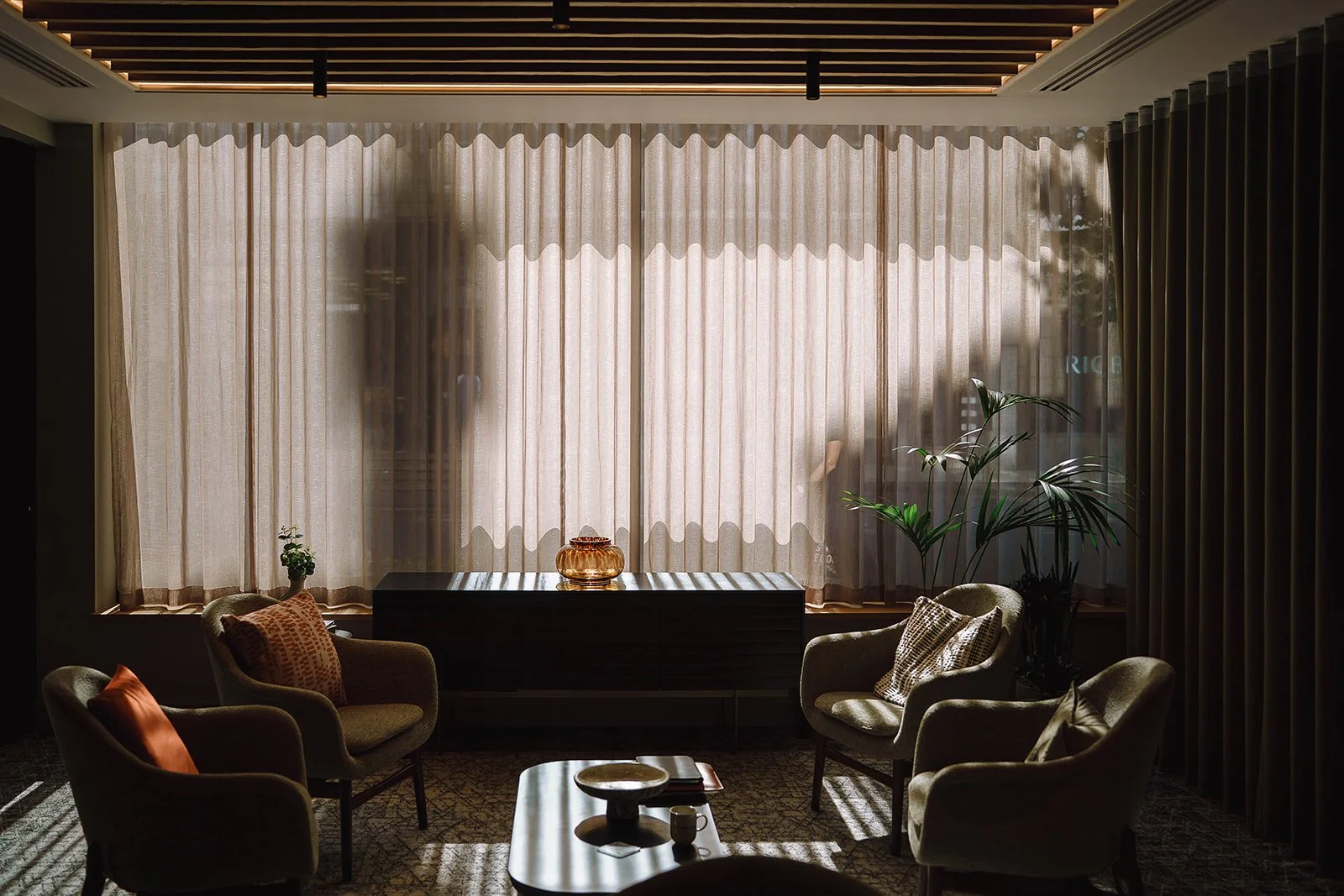 Elegant hotel lounge with armchairs, sheer curtains, wooden credenza, indoor plants, and a coffee table