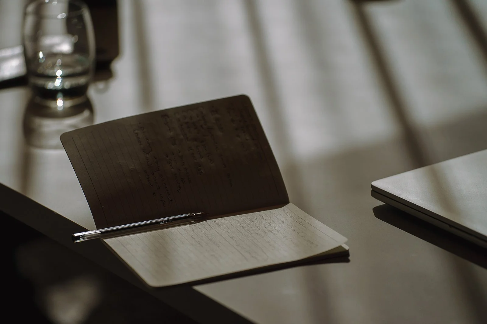 Open handwritten notebook with a pen beside a glass of water and closed laptop on a desk