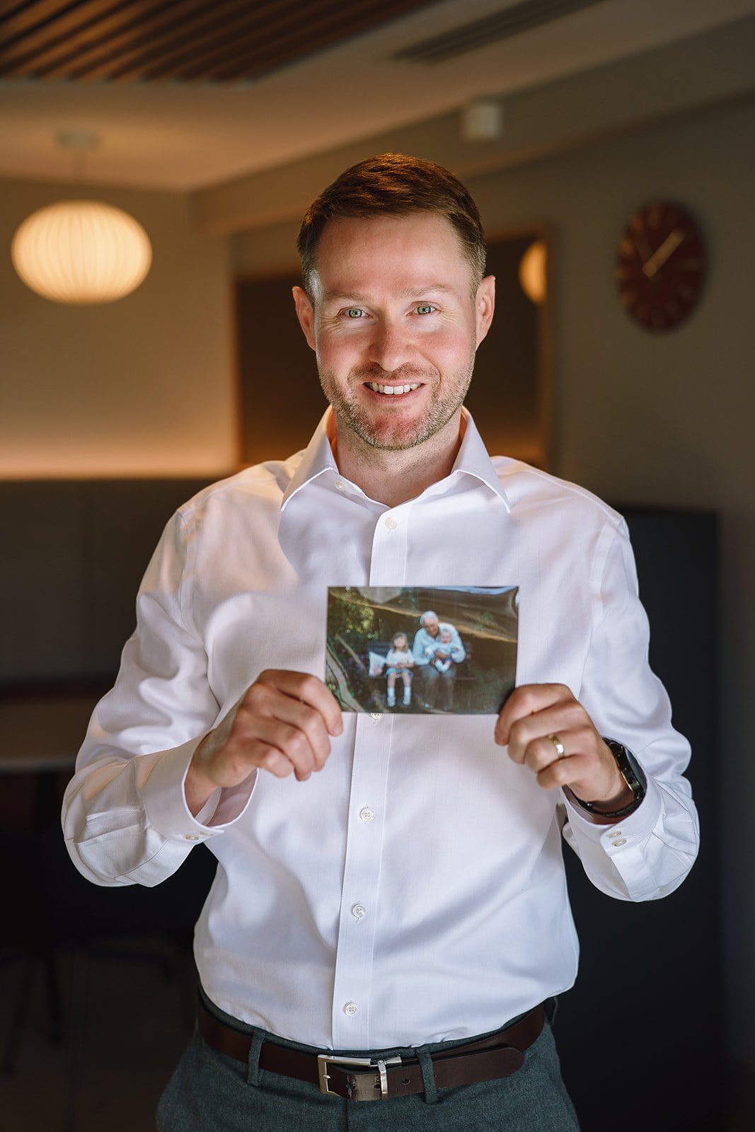Smiling man in a white dress shirt holding a photograph of an older man and child indoors