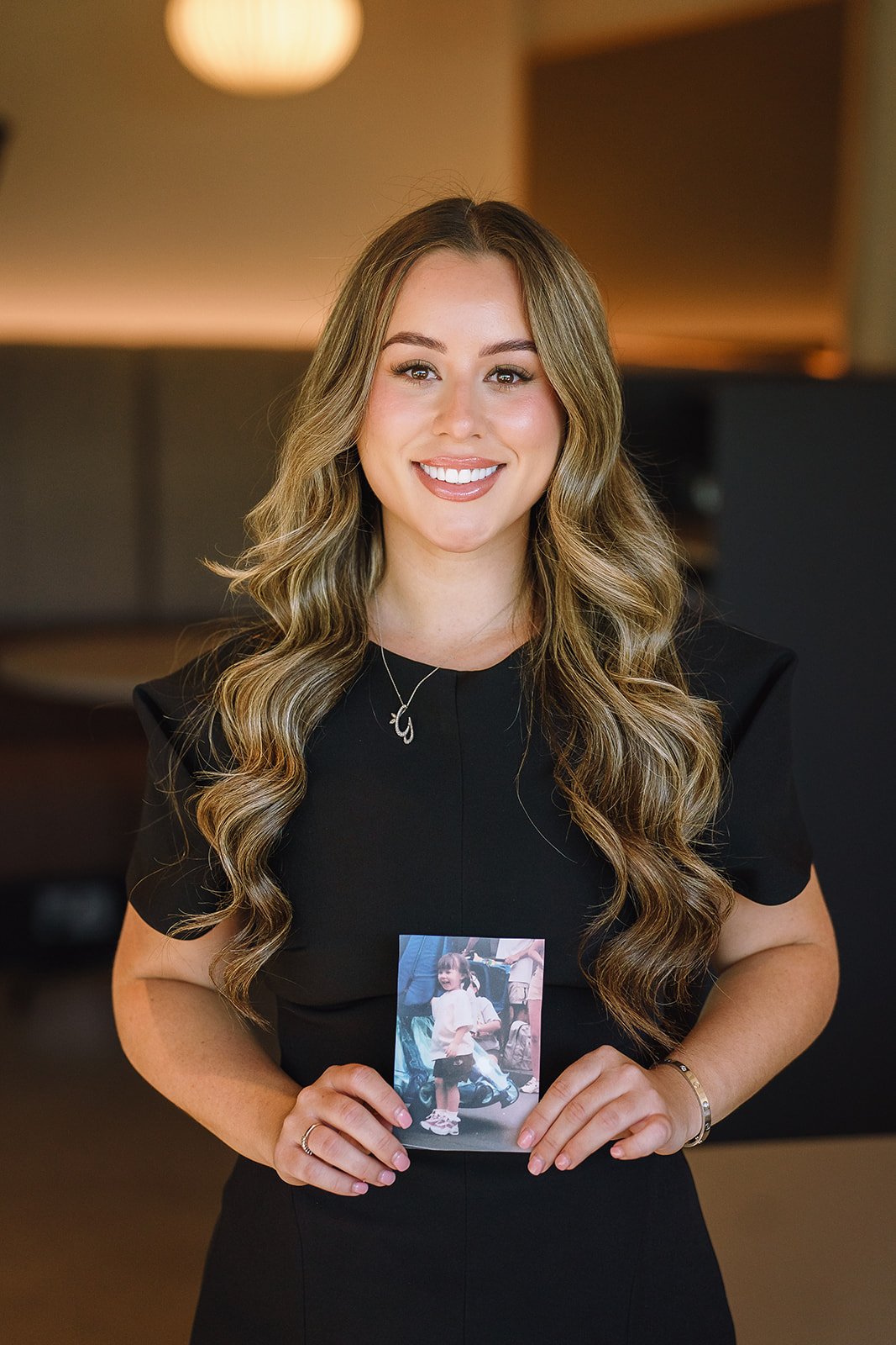 Smiling woman with long wavy hair holding a childhood photo while wearing a black dress.