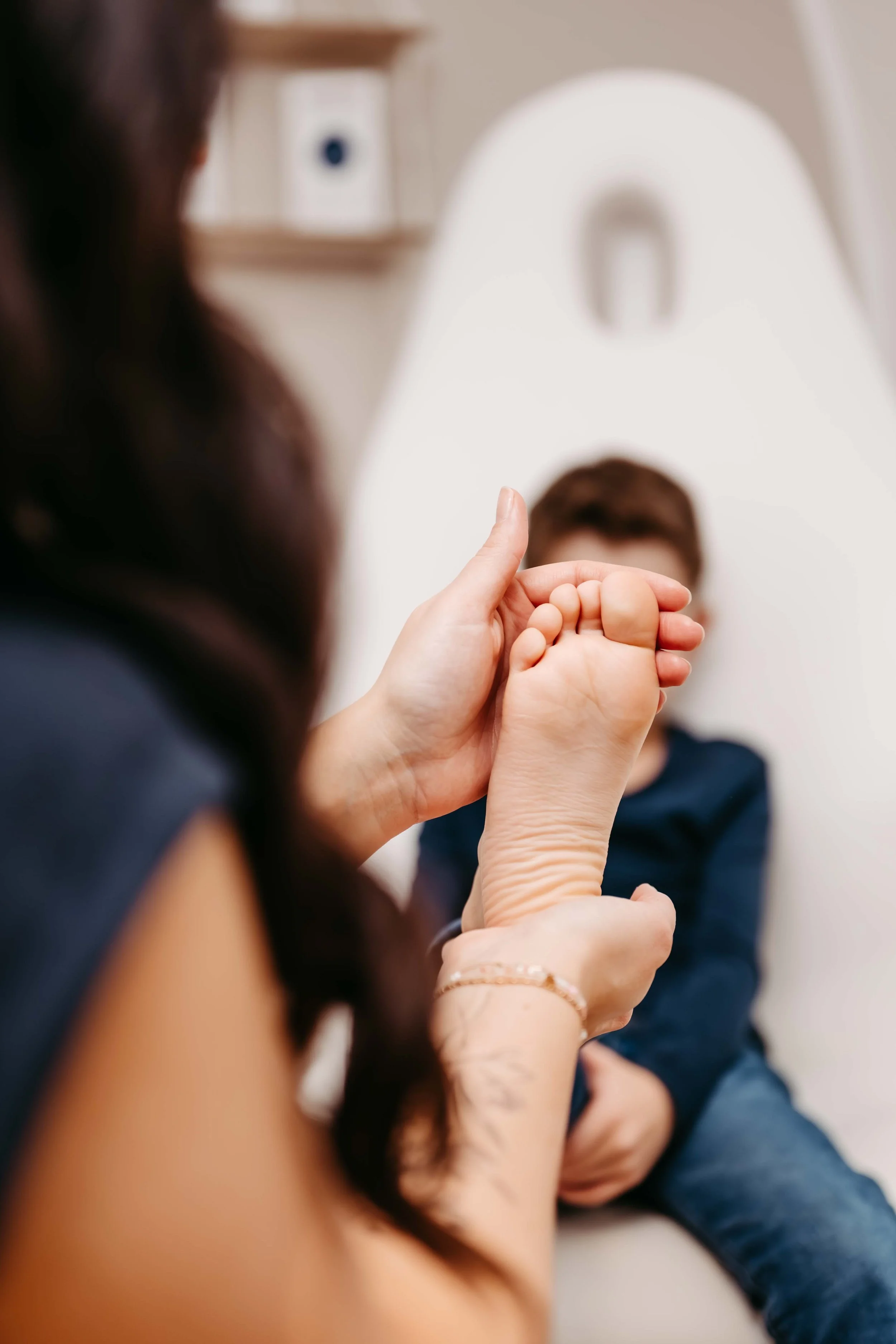 Eine Erwachsene Person hält die Füße eines Kindes, das vor ihr sitzt, in den Händen. Das Kind sitzt auf einem weißen Stuhl und ist im Hintergrund sichtbar.