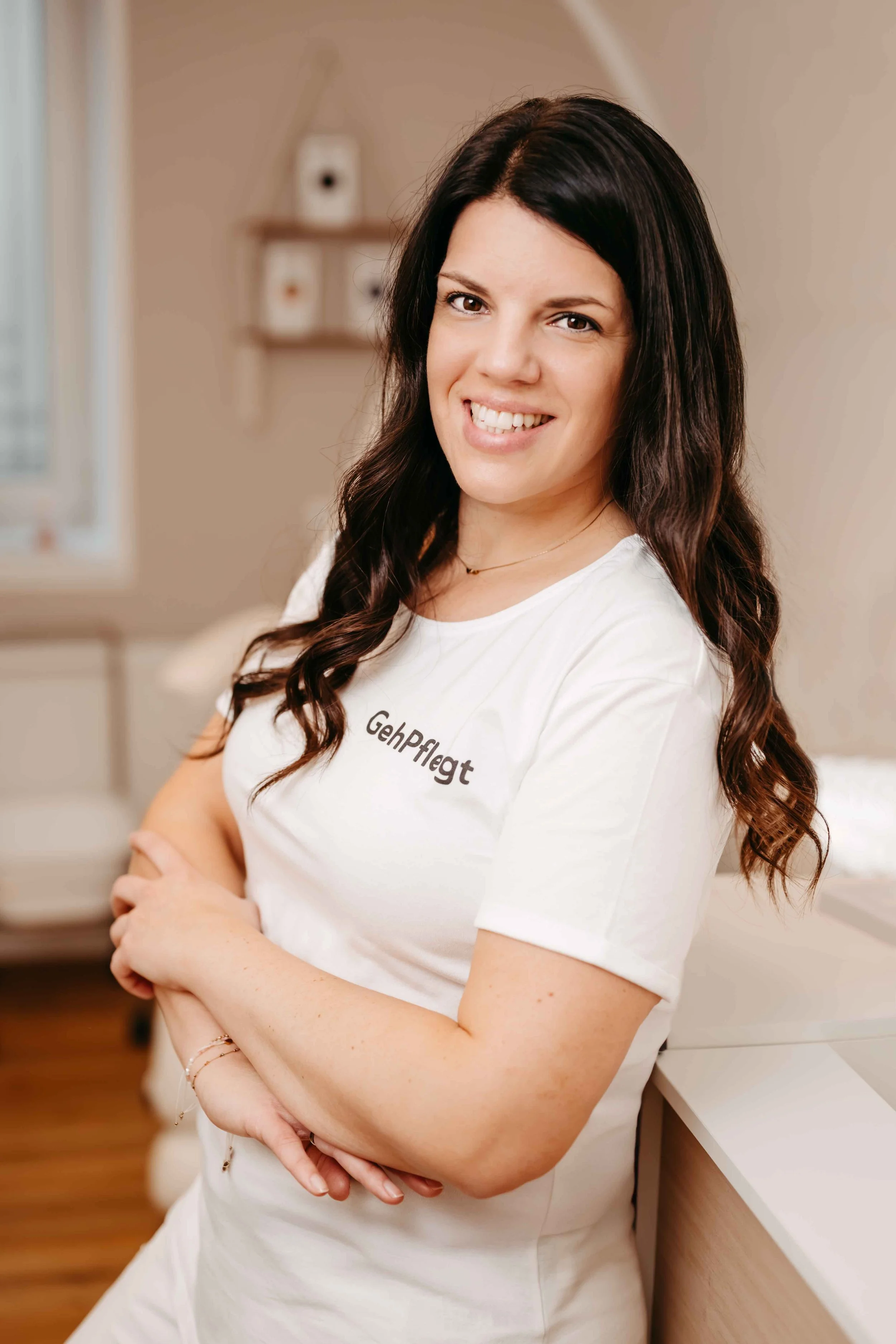 Eine Frau mit schwarzen, gewellten Haaren, trägt ein weißes T-Shirt mit dem Schriftzug "GehpPflegt" und lächelt in die Kamera, während sie die Arme vor der Brust verschränkt.