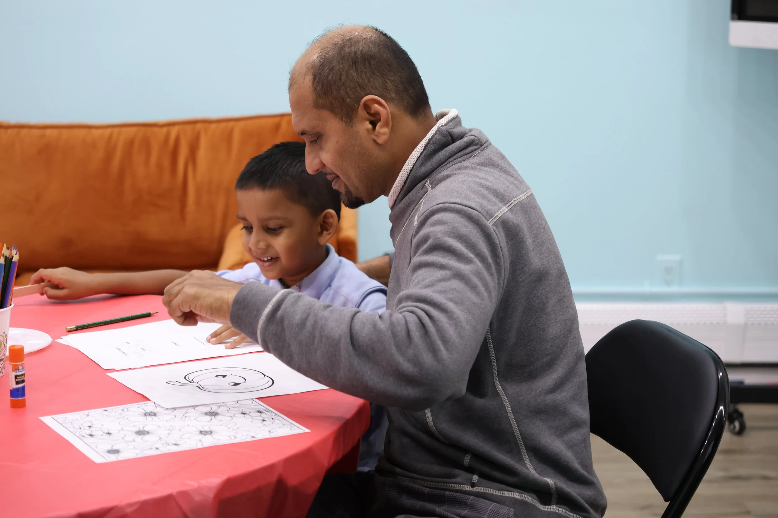 An adult man and a young boy sitting at a table, engaged in a drawing or coloring activity. The boy is smiling, and the man is guiding him, with papers, coloring supplies, and a drawing of a smiling face on the table.