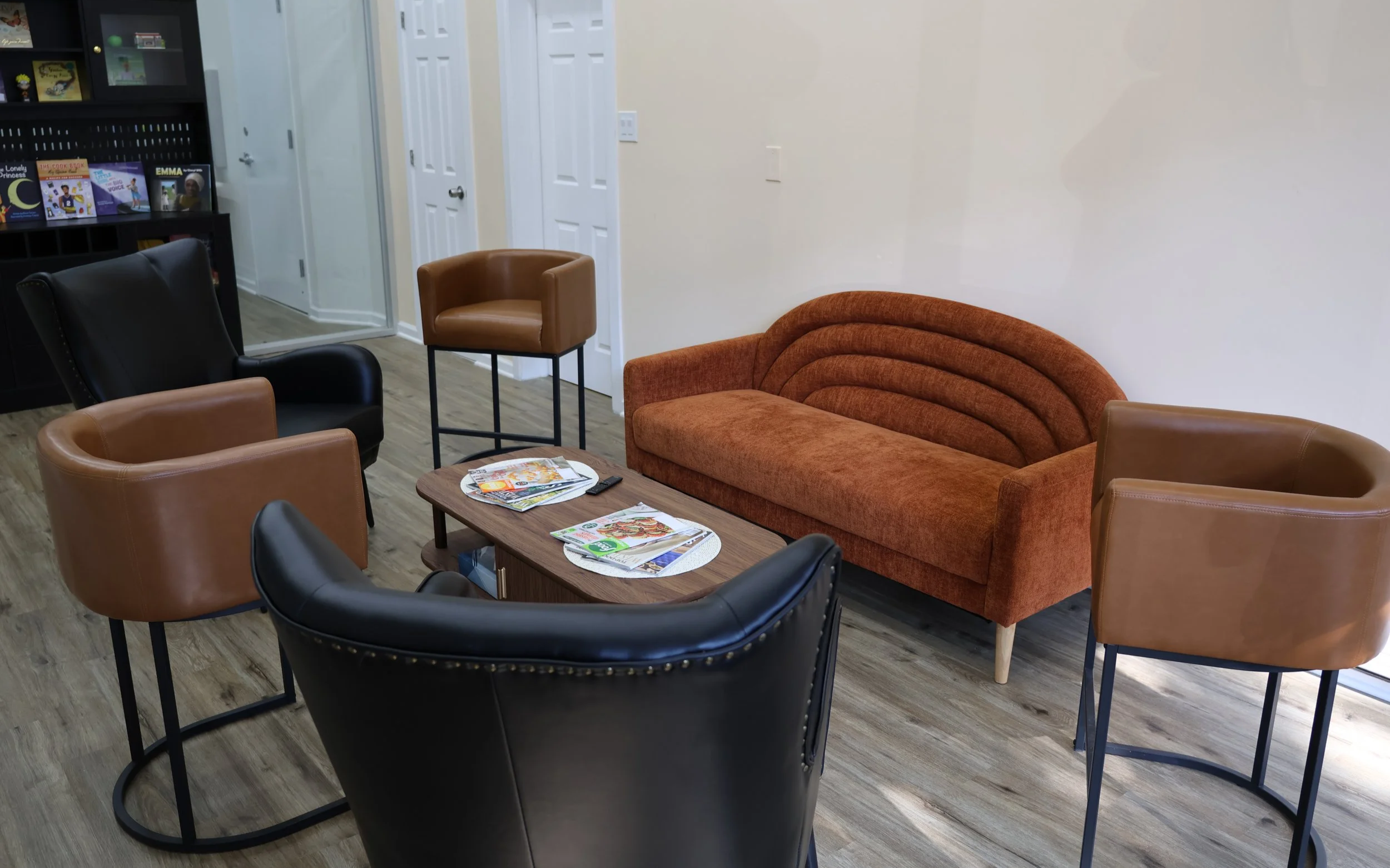 A cozy seating area with a burnt orange mid-century modern sofa, four armchairs in black and brown, and a small wooden coffee table with magazines and a remote control. The room has light-colored walls and wood laminate flooring.