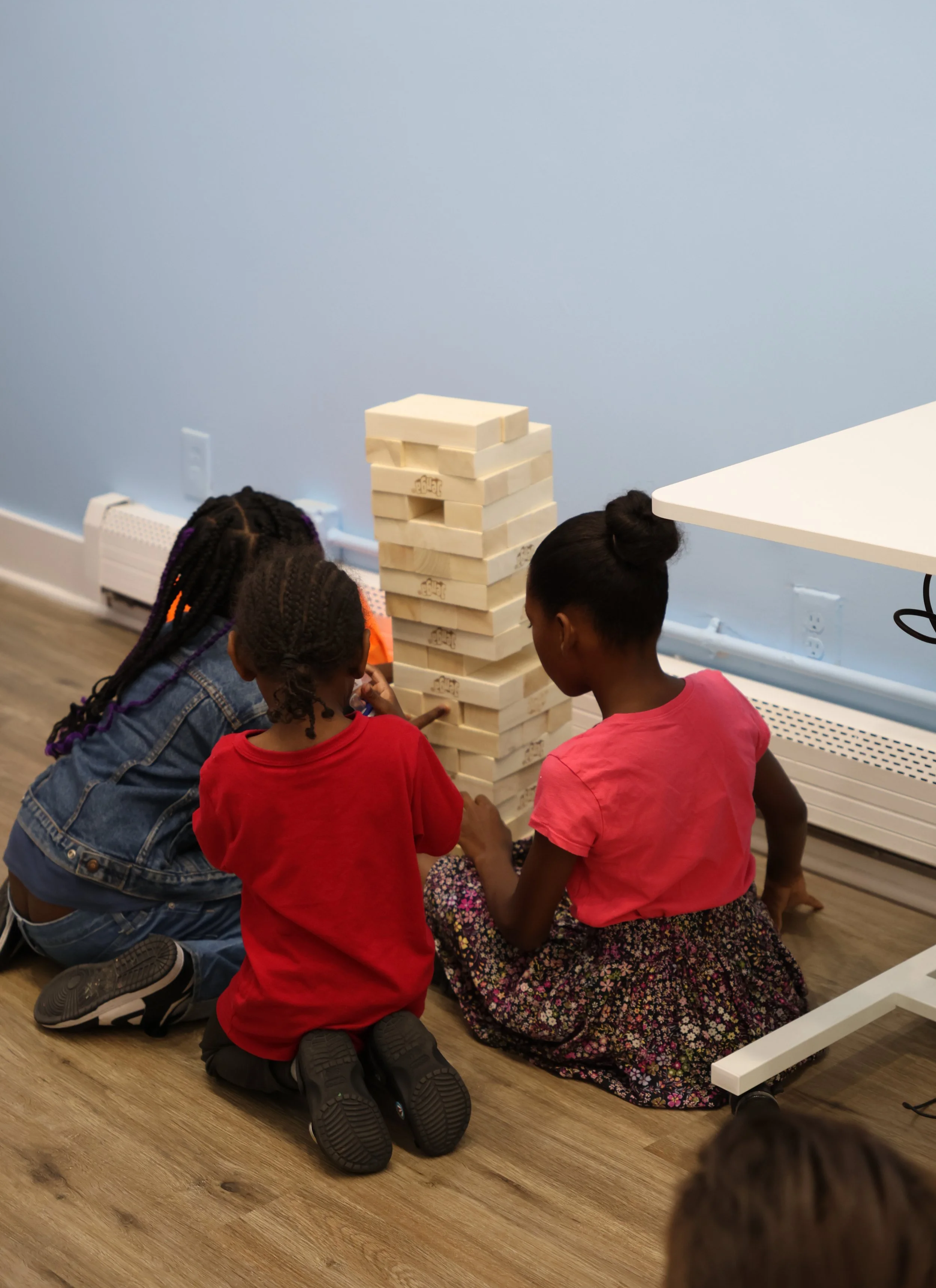 Three children are sitting on the floor, playing a giant Jenga game with large wooden blocks in a room with light blue walls and wood flooring.