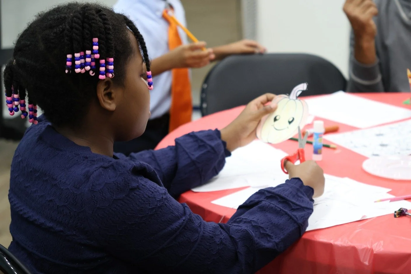 A young girl with braided hair and colorful beads, wearing a navy blue top, sitting at a table with a red tablecloth, holding a paper pumpkin craft with a happy face, scissors, and craft supplies on the table.