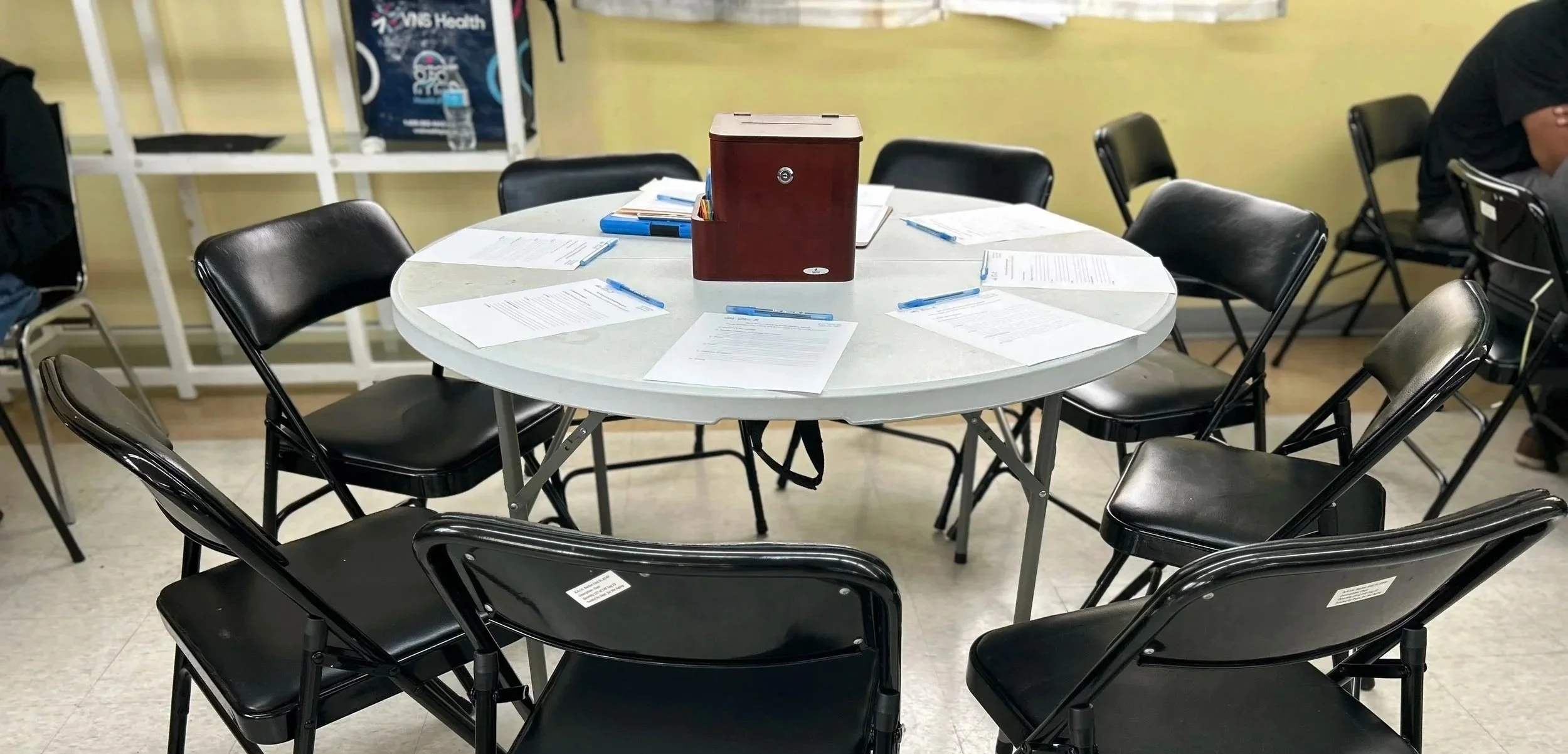 Round table with eight black chairs arranged around it, set with papers and blue pens, with a brown box or container on top of the table in a meeting or classroom setting.
