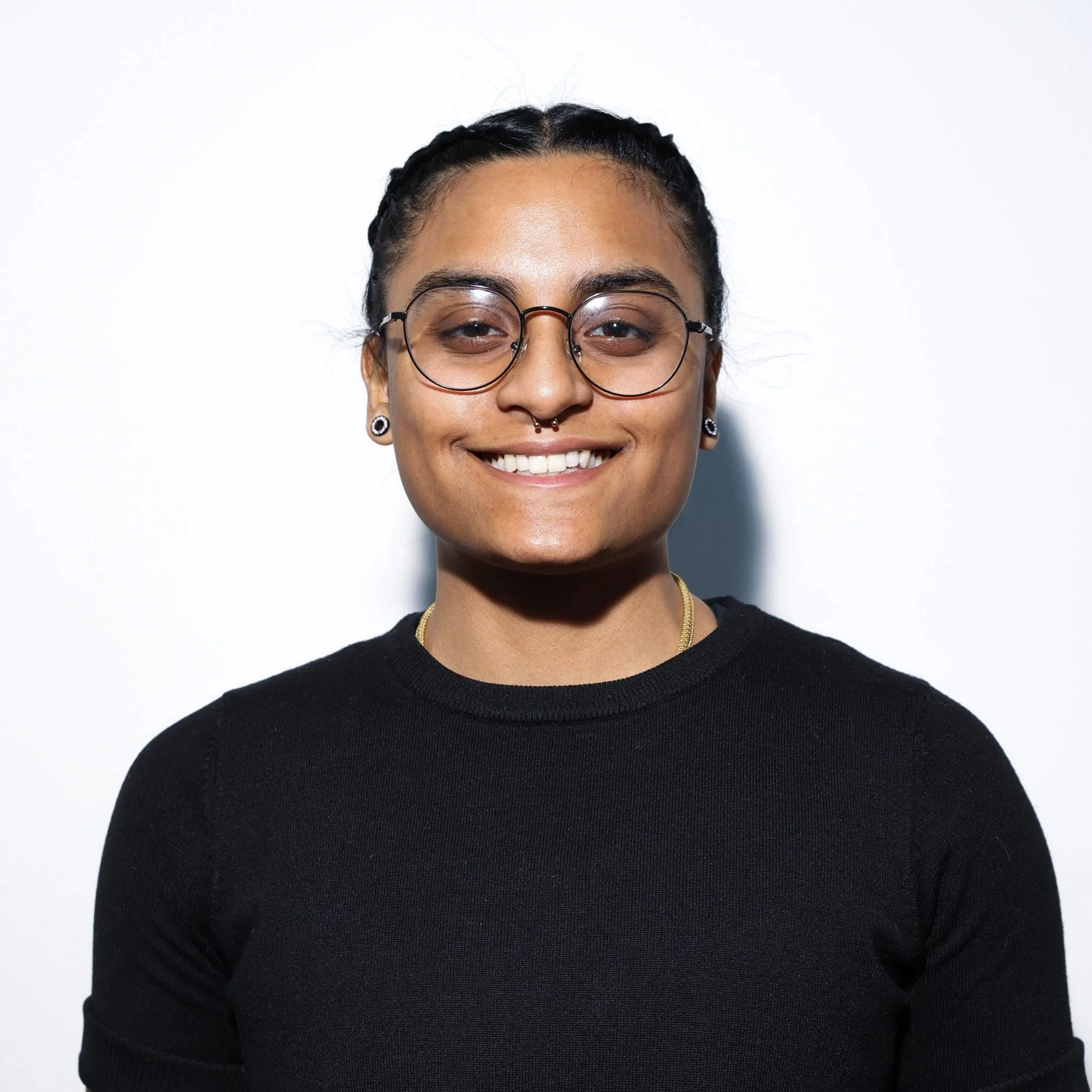Smiling woman with short hair, glasses, septum piercing, wearing a black top, standing against a white background.
