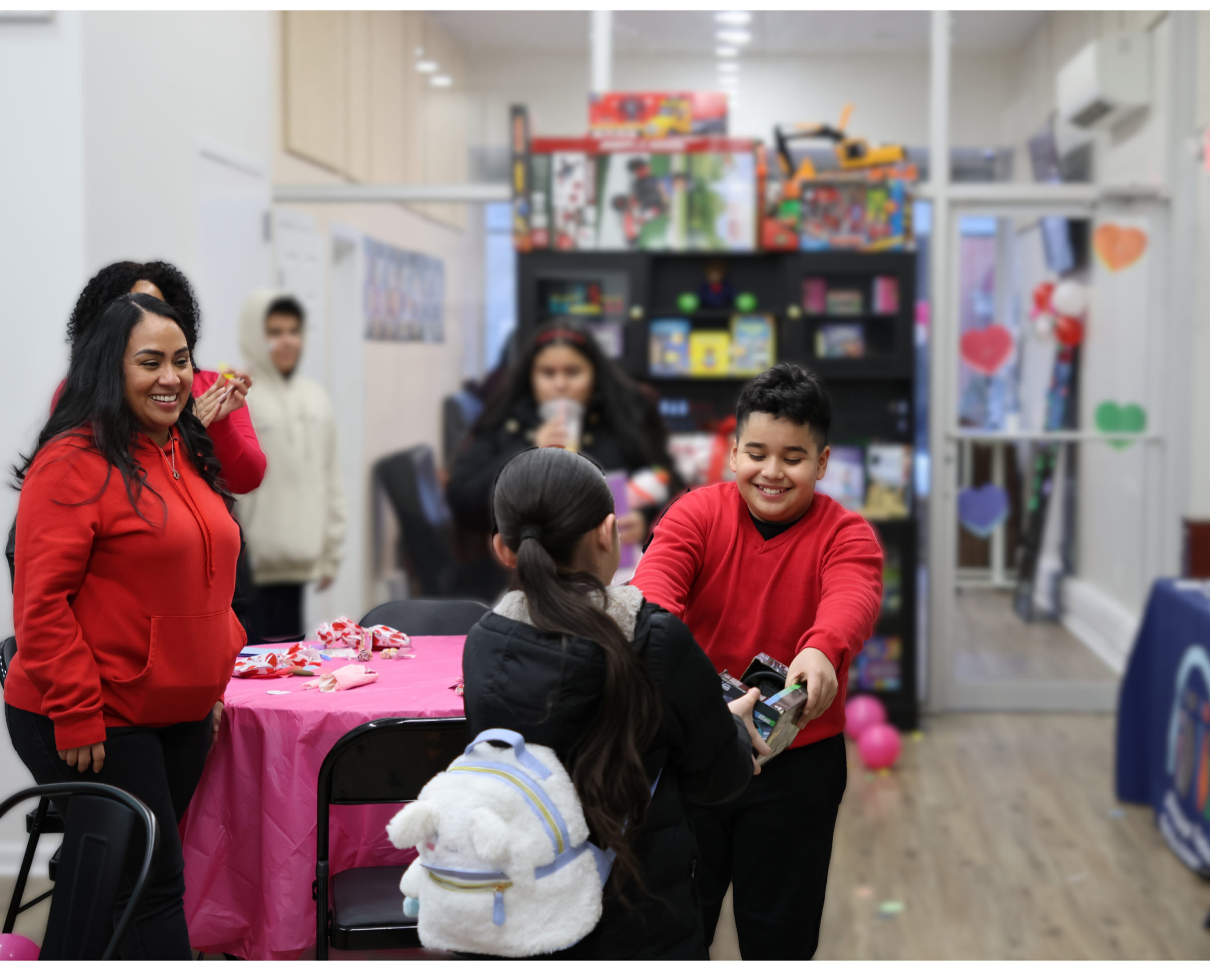 Children and adults celebrating Valentine's Day at a party, exchanging gifts, and surrounded by heart decorations, pink balloons, and candy.