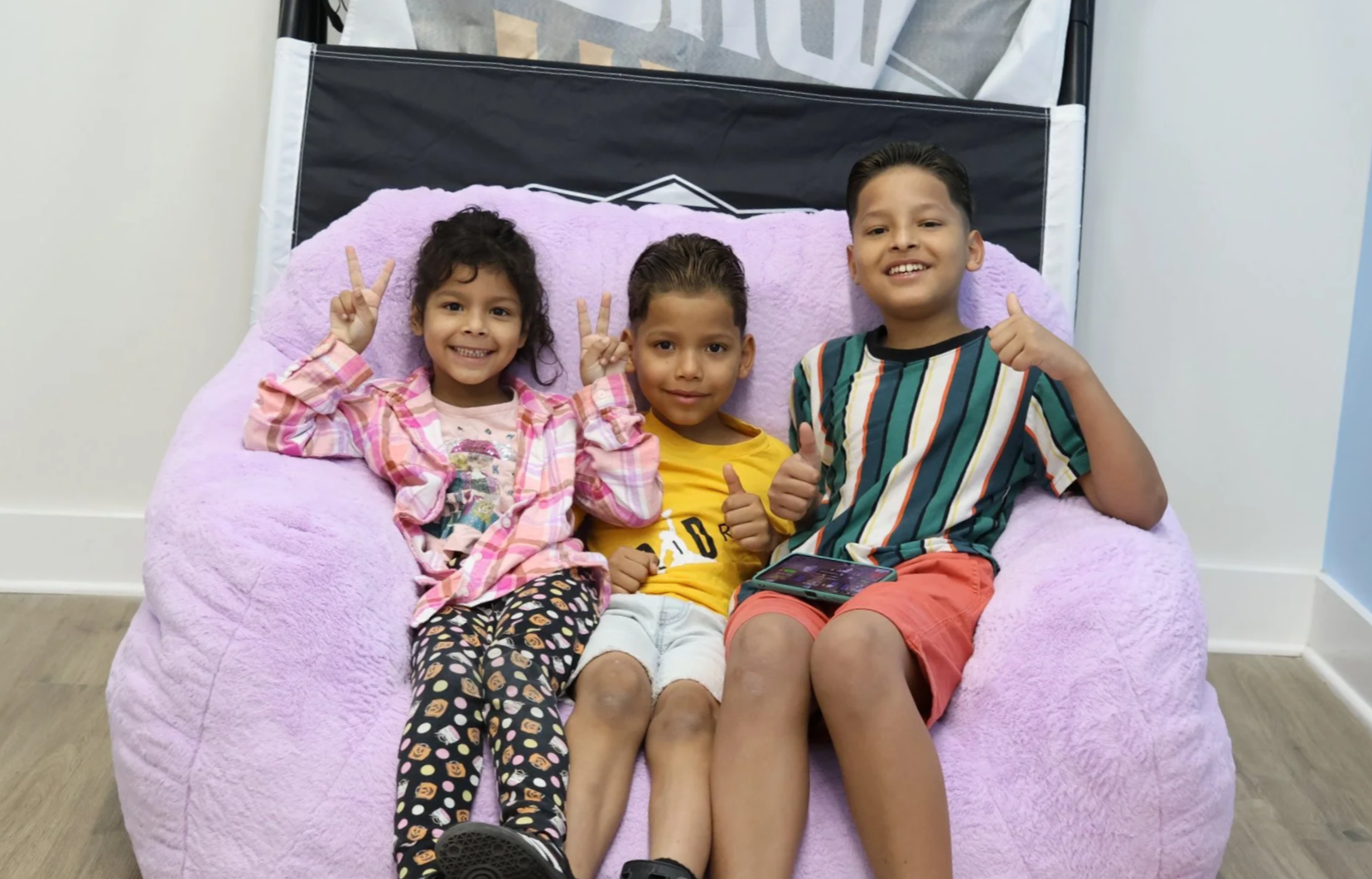 Three children sitting on a large pink plush chair, smiling and making peace signs and thumbs up, in a room with white walls, a blue wall, and wooden flooring.
