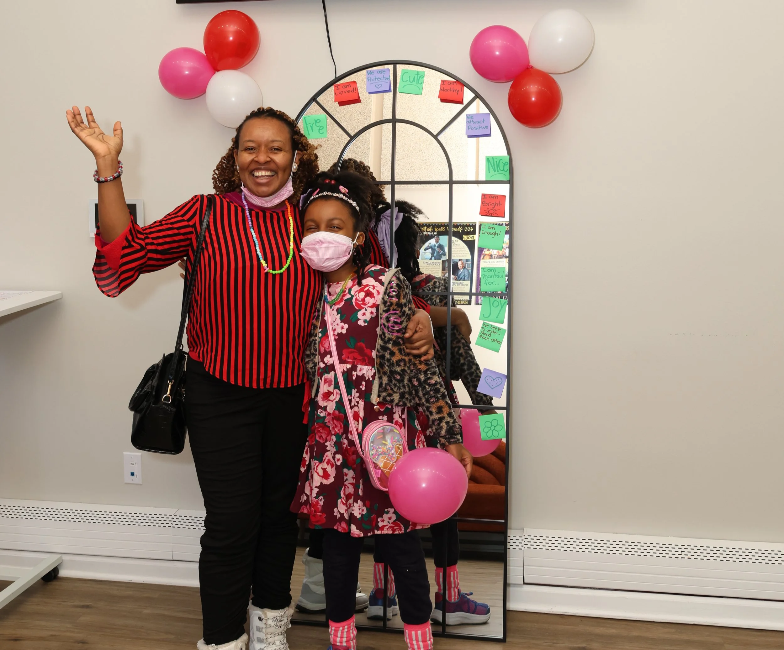 A woman and a girl smiling and waving at the camera, standing indoors in front of a decorative mirror with colorful notes and balloons around it. The woman has curly hair and is wearing a red and black striped blouse, and the girl has braids, a pink face mask, a floral dress, and is holding pink balloons.