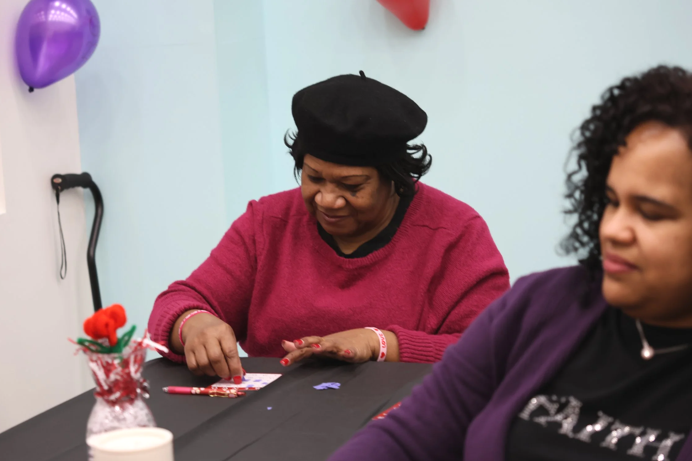 Two women sit at a table decorated with Valentine's Day items, crafting or decorating cards with love-themed designs.