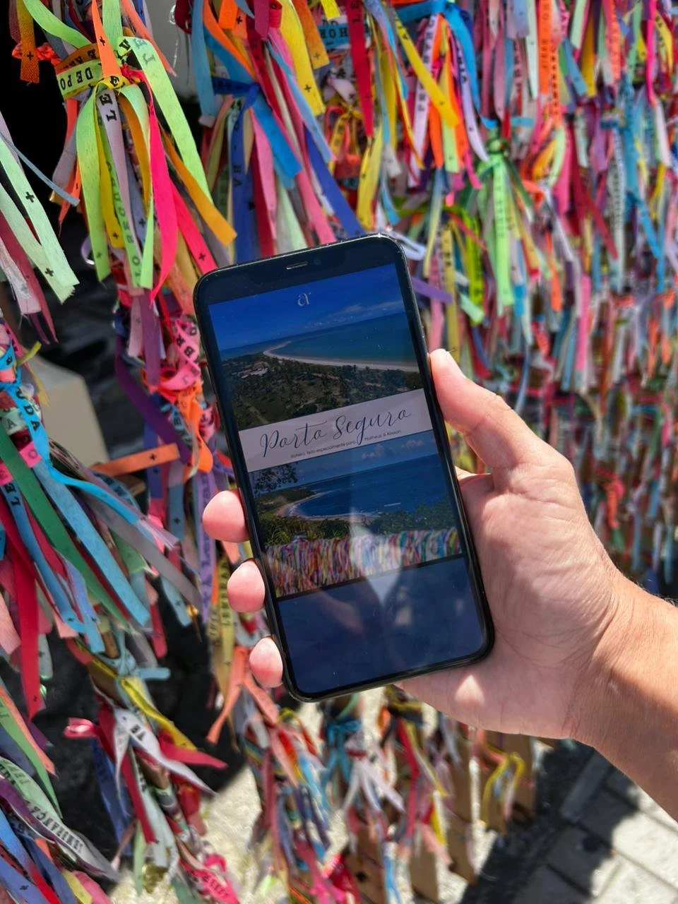Pessoa segurando um celular com o roteiro digital de Porto Seguro na frente de um painel com pulseiras do senhor do Bonfim