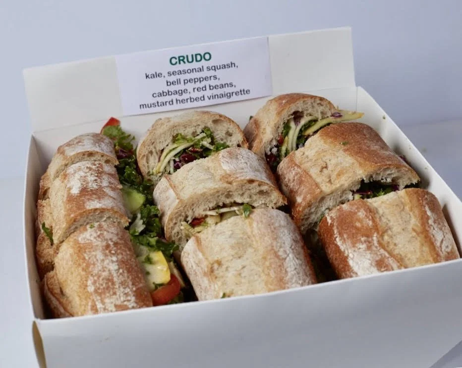 A white box containing six sandwiches with cut bread, filled with vegetables and greens, and a label with ingredients like kale, squash, peppers, cabbage, beans, and vinaigrette.