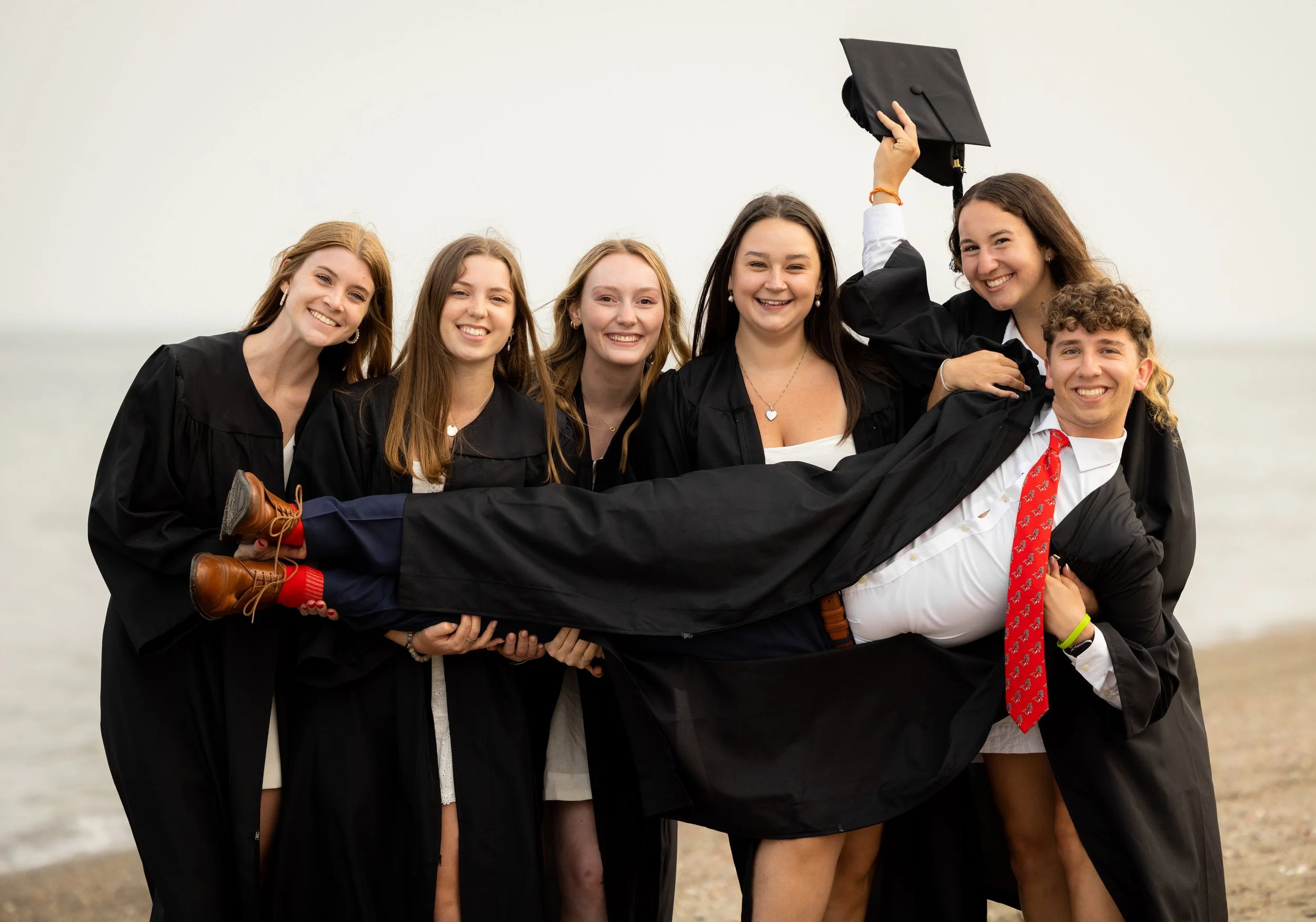 a beach graduation shoot with six graduates, holding one of them sideways as they all pose for a photo