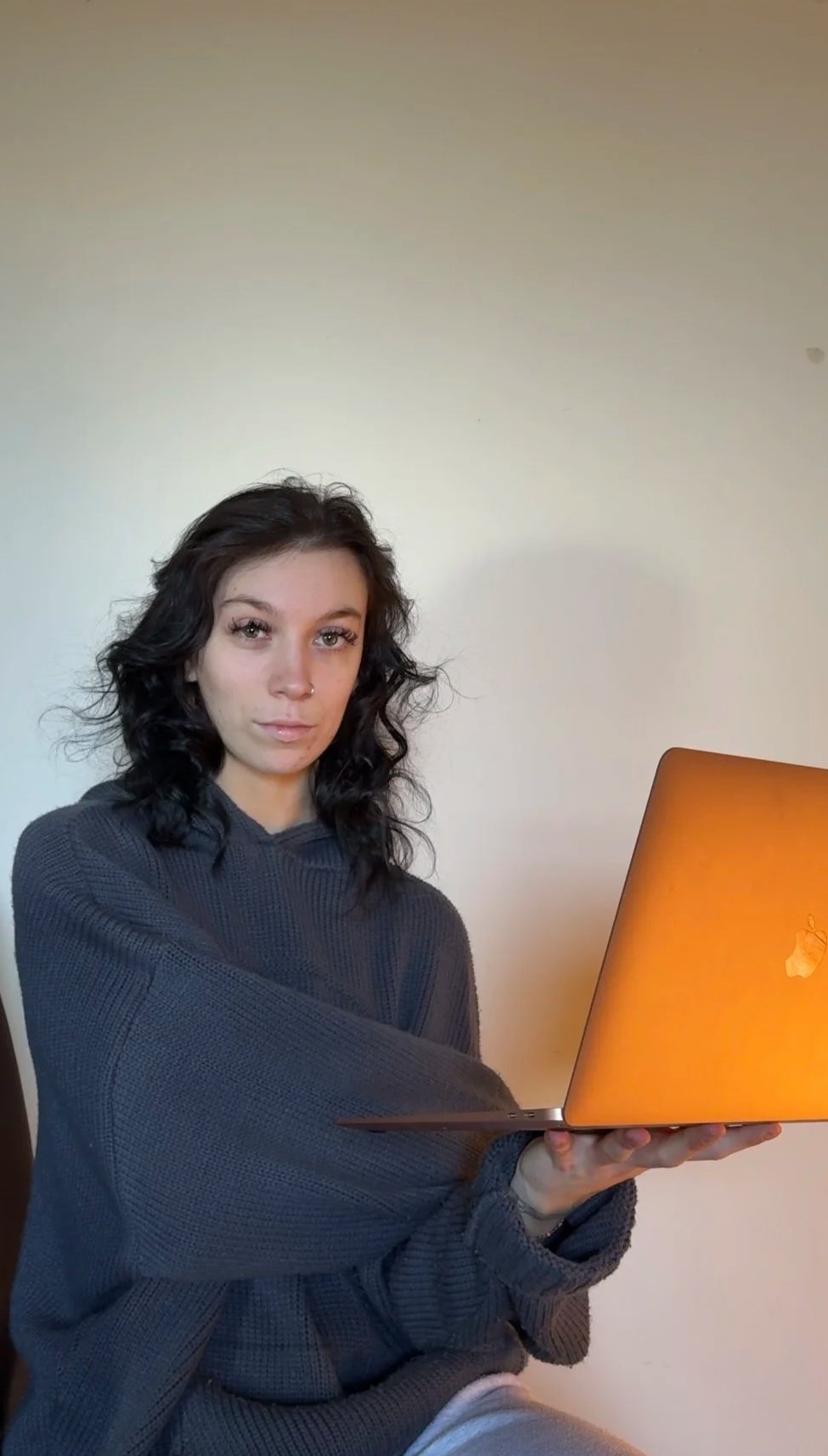 A young woman with dark, wavy hair and light skin is holding an orange laptop. She is wearing a dark gray, long-sleeve sweater and is sitting against a plain wall.