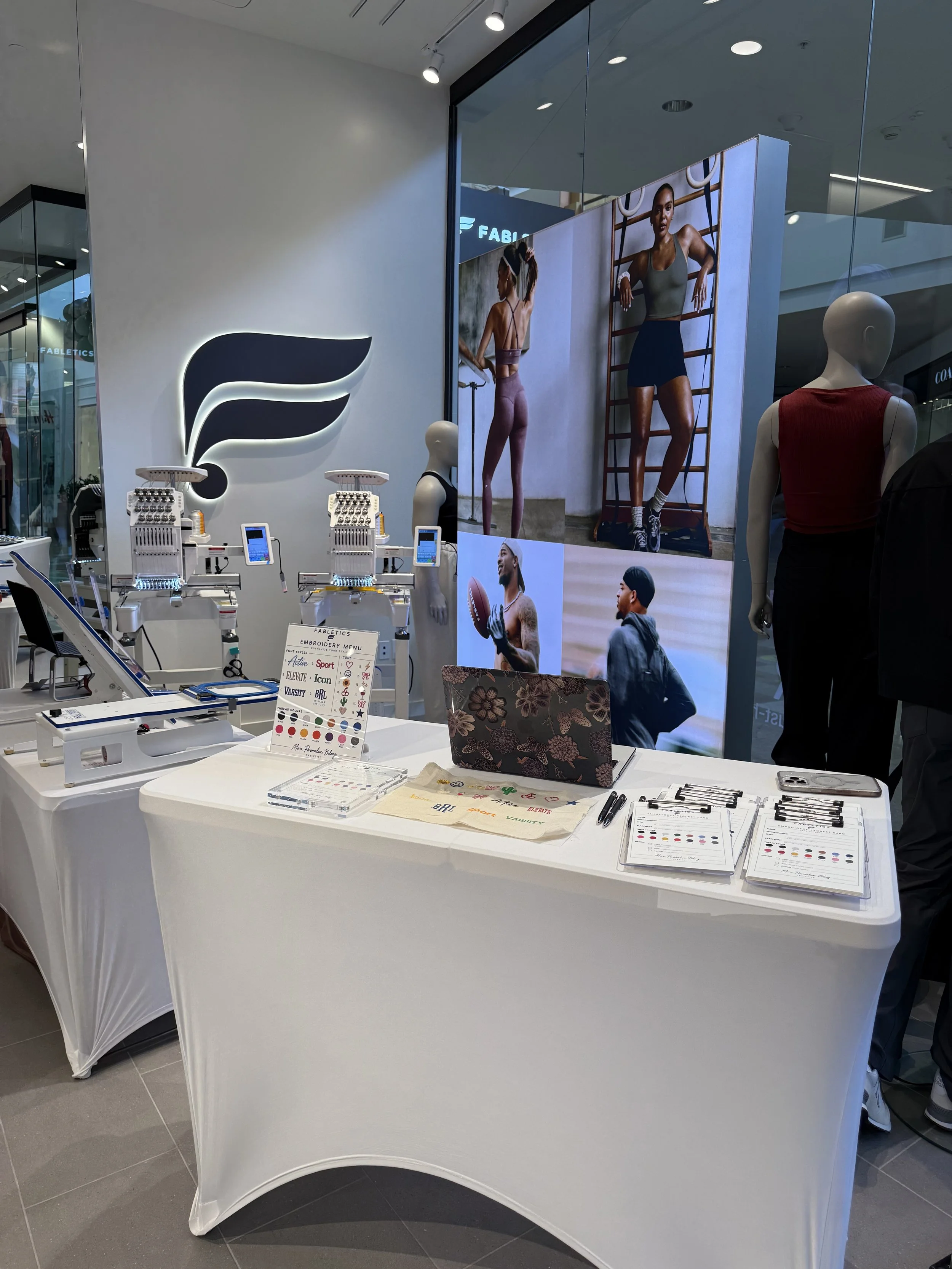 Display table with embroidery supplies including thread, needles, and color charts in a retail store. Mannequins and large posters of athletic models are visible in the background.