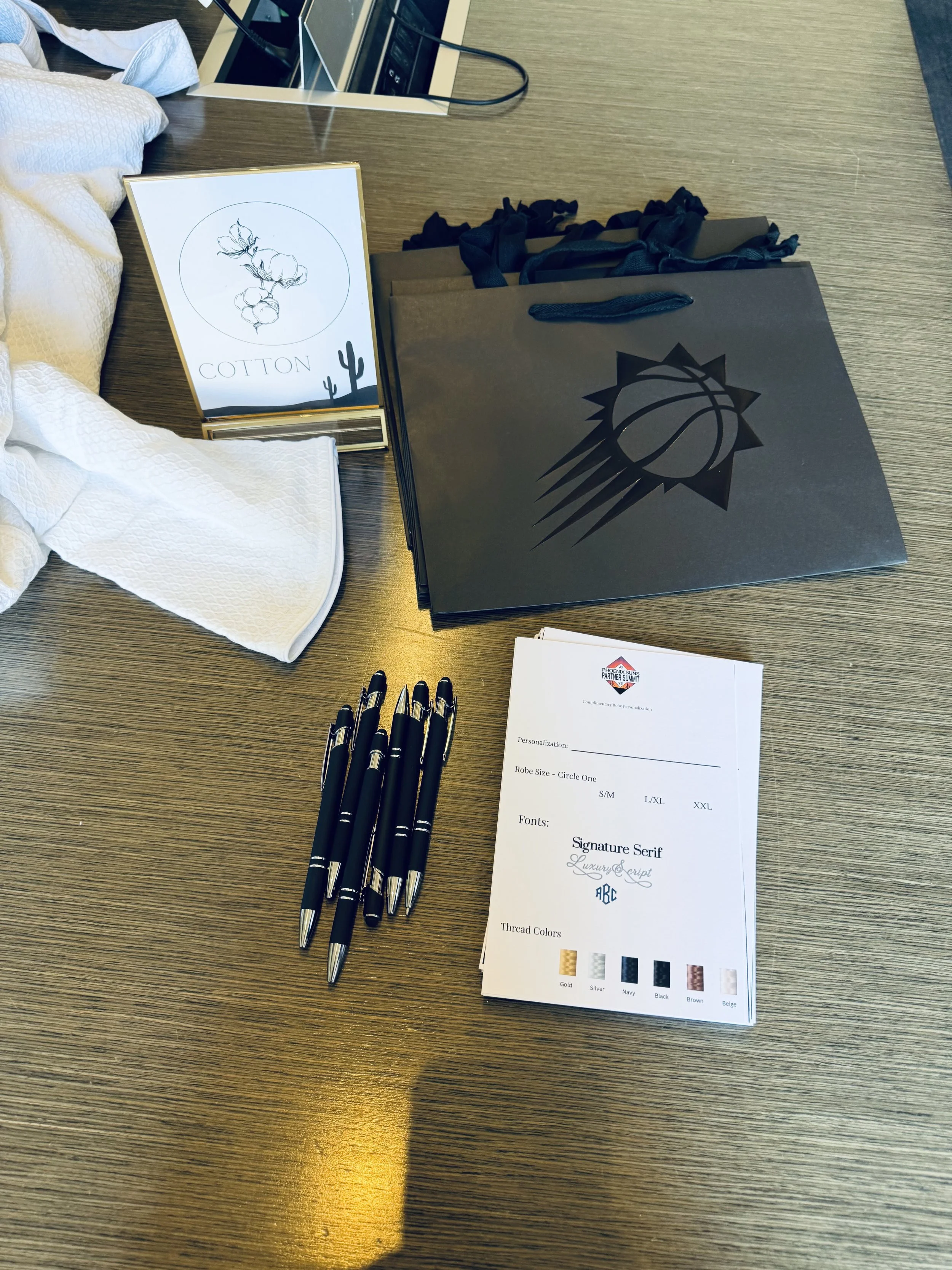 On a wooden table, there are several black pens, a stack of white papers with font and color options for customization, a black shopping bag with a basketball logo, a white cloth or towel, and a sign with the word 'COTTON' and a sketch of cotton plant with desert cacti in the corners.