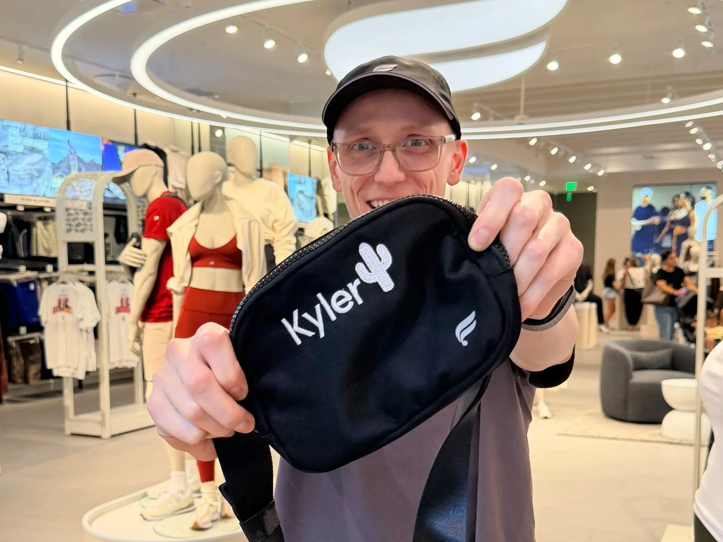 A man with glasses, wearing a cap, smiling, holding a black Kyler bag inside a retail store with mannequins dressed in athletic clothing and computer screens in the background.